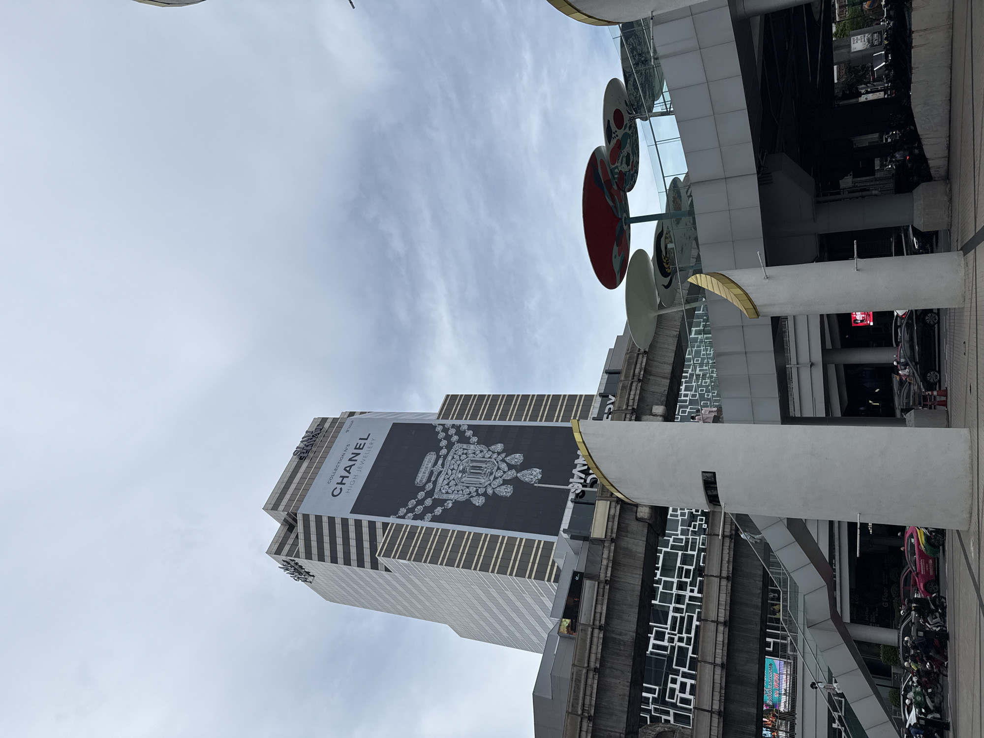 The image, taken from a low angle, shows the skyline of Bangkok, Thailand. A tall, modern building is prominently featured, adorned with a large advertisement for "CHANEL HIGH JEWELLERY". The building itself is a mixture of white and striped gray tones. To the right of the building, an elevated structure, potentially a skywalk or pedestrian bridge, is visible with repetitive square patterned designs. The sky above is overcast with gray and white clouds. An artistic installation consisting of circular discs is also partially visible on the right side. The scene seems to be taken from a busy urban environment with hints of infrastructure, such as road or pedestrian pathways, and parked motorcycles and other vehicles in the periphery.