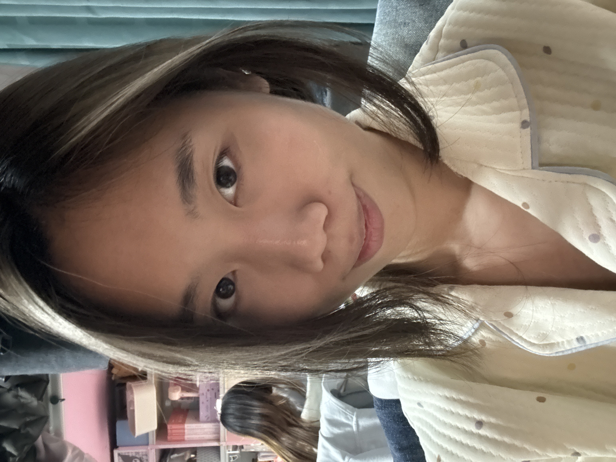 A close-up, angled selfie of an Asian woman with medium-length brown hair and blonde highlights. She is wearing a light-colored, quilted pajama top with small, dark polka dots and a gray trim. The background includes a blurred view of a person with long dark hair wearing a light-colored shirt and denim, a shelf with books and small items, and a light pink wall, suggestive of a domestic interior setting. This photo doesn't allow for the conclusion that the image was taken in the city of Nan'an District, China.