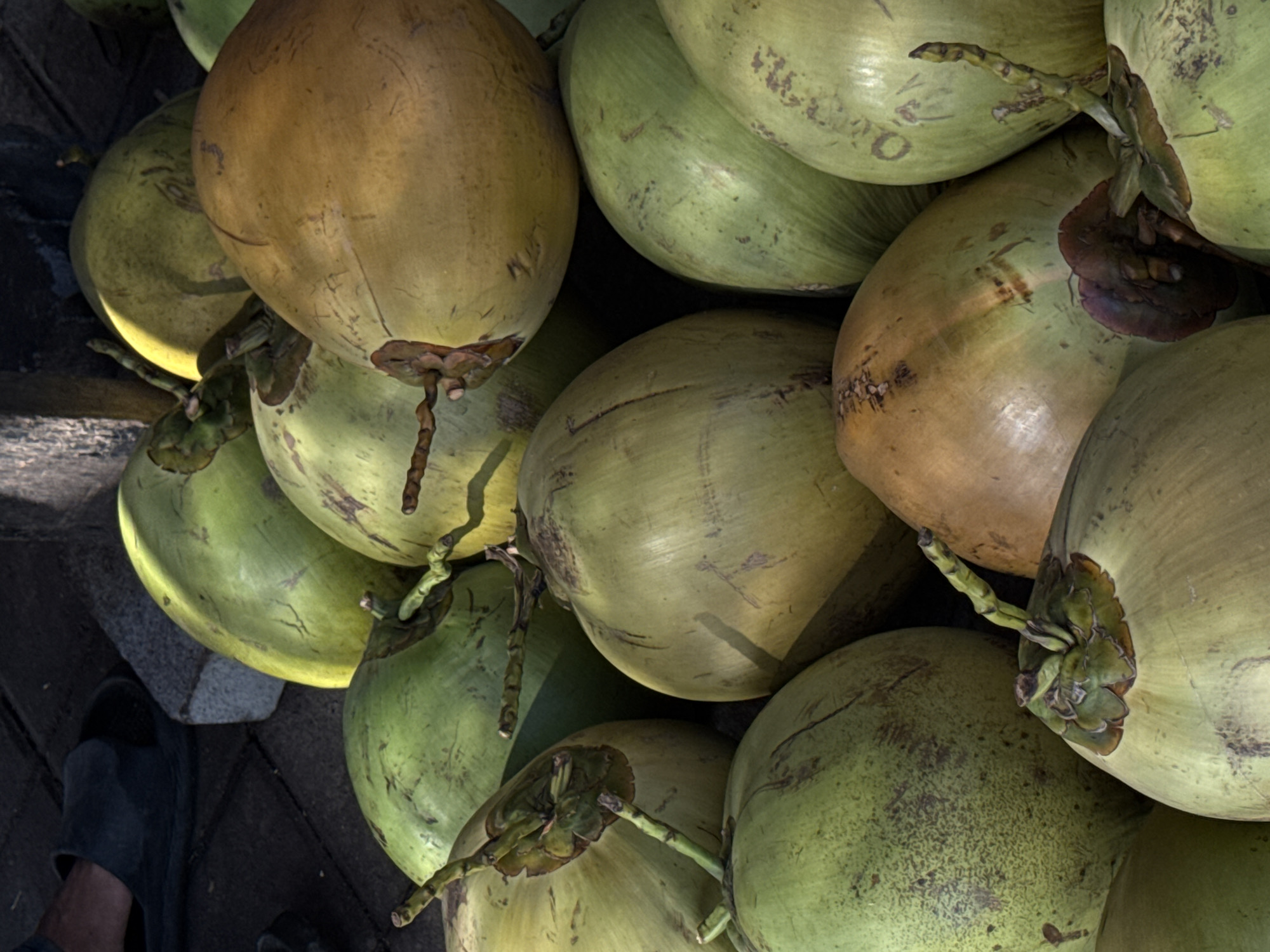 The image presents a close-up view of a pile of young coconuts. Most of the coconuts are green with varying shades and some brown. Some have stems, while others appear to have been freshly cut from the tree. The coconuts are piled together, obscuring a clear view of the surface they rest on. A pair of black shoes/slippers is visible in the lower-left corner. The specific location within Unknown, Indonesia, cannot be determined from this image.