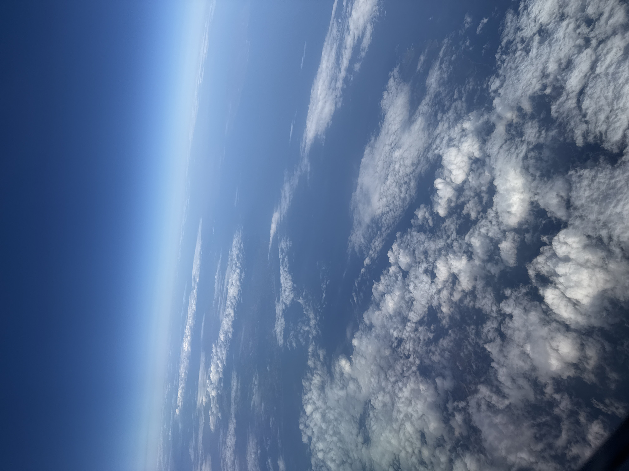 The image is an aerial view of clouds and ocean under a clear blue sky. The perspective is from a high altitude, likely from an airplane, looking down at the Earth's surface. The lower part of the image is filled with fluffy white clouds, which transition into a vast expanse of blue ocean. In the distance, the ocean meets the sky at the horizon, with a gradient of lighter blue to a deeper blue as the sky extends upwards. There are no visible man-made structures or indications of specific locations within the city of Νεοχώρι, Greece. The scene is natural and serene, dominated by the elements of sky and sea.