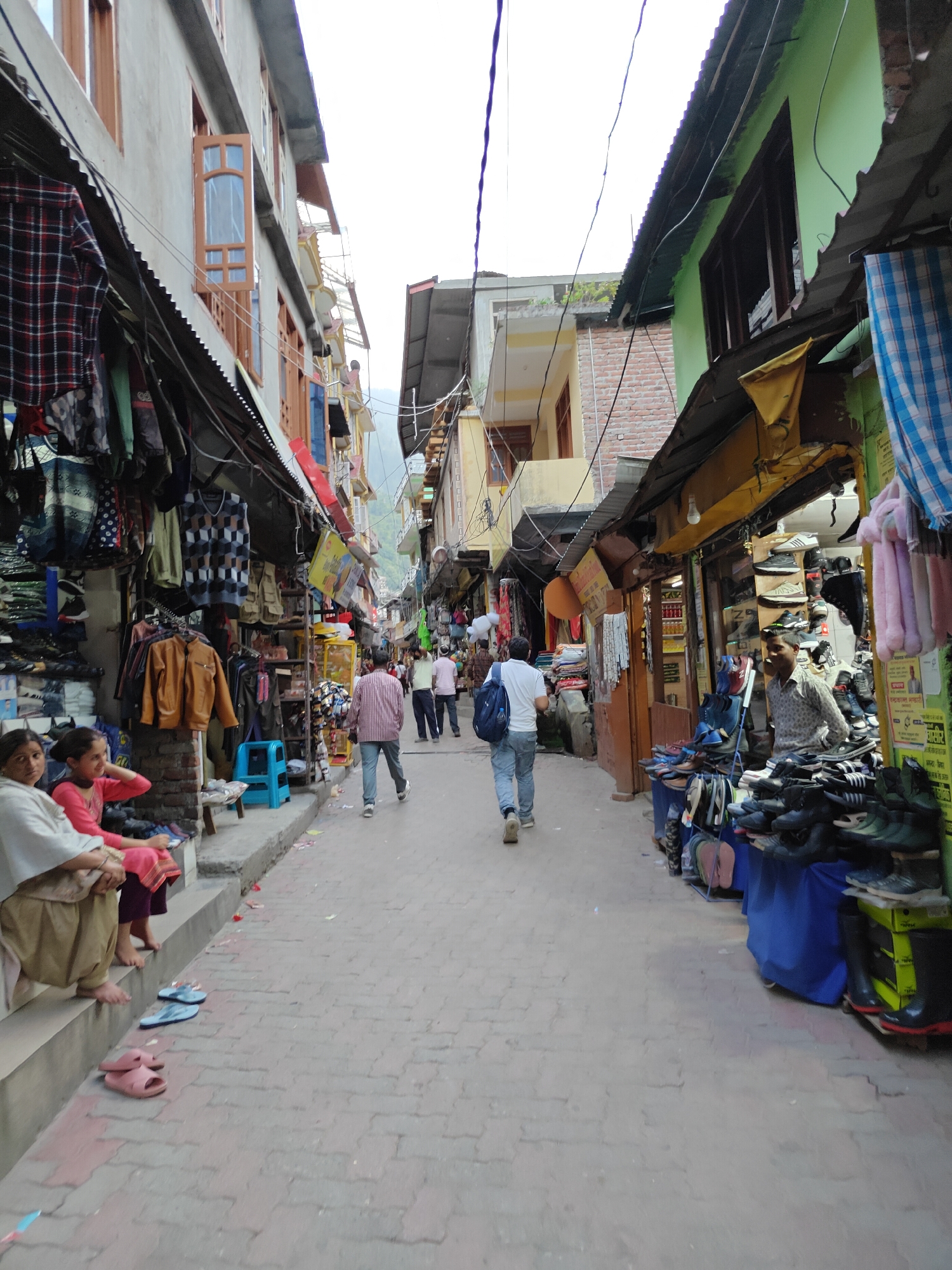 The image presents a vibrant, narrow market street in Manikaran, India, bustling with activity under an overcast sky. The street, paved with interlocking bricks, is flanked by multi-story buildings housing numerous small shops.
On the left side, stores extend outward with corrugated metal awnings, displaying a variety of clothing including plaid shirts, sweaters, and leather-look jackets. In the foreground, two women and a young girl are seated barefoot on a low concrete ledge, observing the street scene. The older woman wears a light-colored shawl and beige pants, while the girl is dressed in pink. Two pairs of sandals, one light blue and one pink, rest on the pavement beside them.
The right side of the street is similarly lined with shops, prominently featuring a footwear stall where a vendor is visible among piles of shoes, including sneakers, sandals, and boots, arranged on tables and the ground, partially draped with a blue tablecloth. Further down, other shops with various merchandise are discernible.
Several people are walking along the street, navigating through the market. A man in a striped shirt and jeans, followed by others, walks towards the background, while another man with a white t-shirt and blue backpack is seen on the right. Power lines crisscross above the street, connecting the buildings. The architecture is characteristic of the region, with various painted facades (light grey, light green, yellow) and prominent wooden window frames, some open.
The bright, overcast sky indicates daytime with soft, diffuse lighting, without harsh shadows. The scene conveys a lively, everyday market atmosphere in what appears to be a hilly or mountainous region. Text on distant signs is not clearly legible.