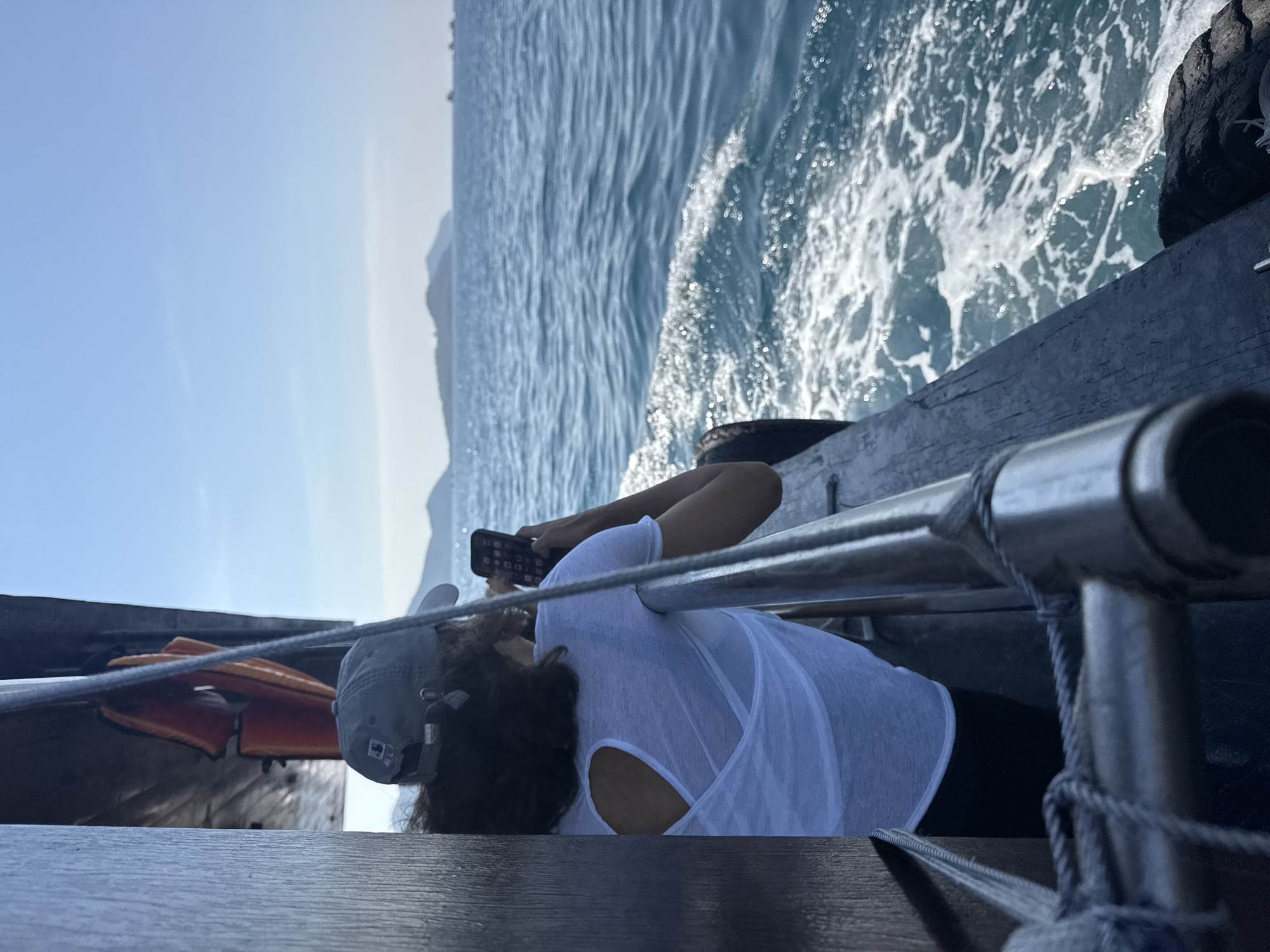 A person wearing a grey baseball cap and a white sheer shirt with a cutout back is standing on the side of a wooden boat. The person is holding a smartphone and appears to be taking a photo or video of the ocean. The boat is moving through the water, creating a wake. The sea is a deep blue, and the sky is clear with some distant islands visible. The scene is set in the waters off the coast of Nam Nha Trang Ward, Vietnam.