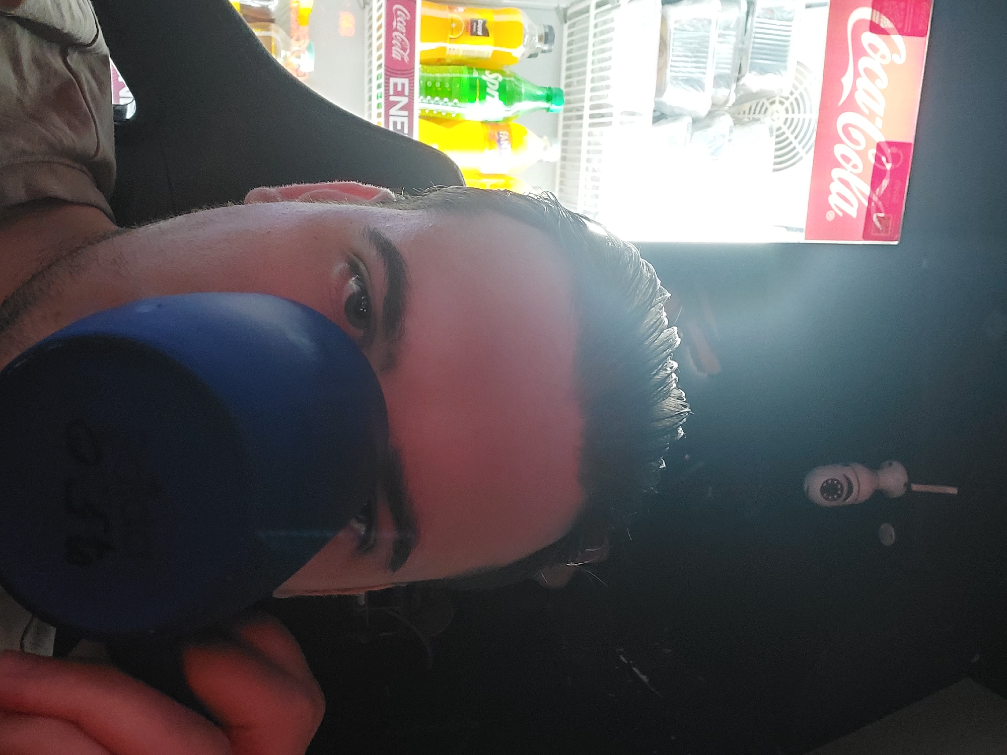 An eye-level indoor shot captures a person partially obscured by a blue cup or bottle, with a brightly lit commercial refrigerator in the background. The person, whose face is largely covered by the blue object, reveals only their eyes, eyebrows, forehead, and part of their nose, looking towards the camera. Their hair appears dark and wet or slicked back. They are wearing a light-colored top and are holding the blue item with their hands, which are partially visible.
In the background, a white commercial refrigerator is brightly illuminated, displaying various bottled beverages. Visible brands include "Coca-Cola" prominently on the side panel, and inside, bottles of "Sprite" (green) and "Fanta" (orange/yellow) are discernible among other yellow-bottled drinks. A part of a fan or cooling vent is visible inside the refrigerator unit. To the right of the person's head, on a dark, flat surface, there is a small, white object resembling a security camera mounted on a stick-like base. The overall lighting suggests a contrast between the person in the foreground and the bright refrigerator in the mid-ground, possibly taken in a convenience store or cafe setting in Tashkent, Uzbekistan.