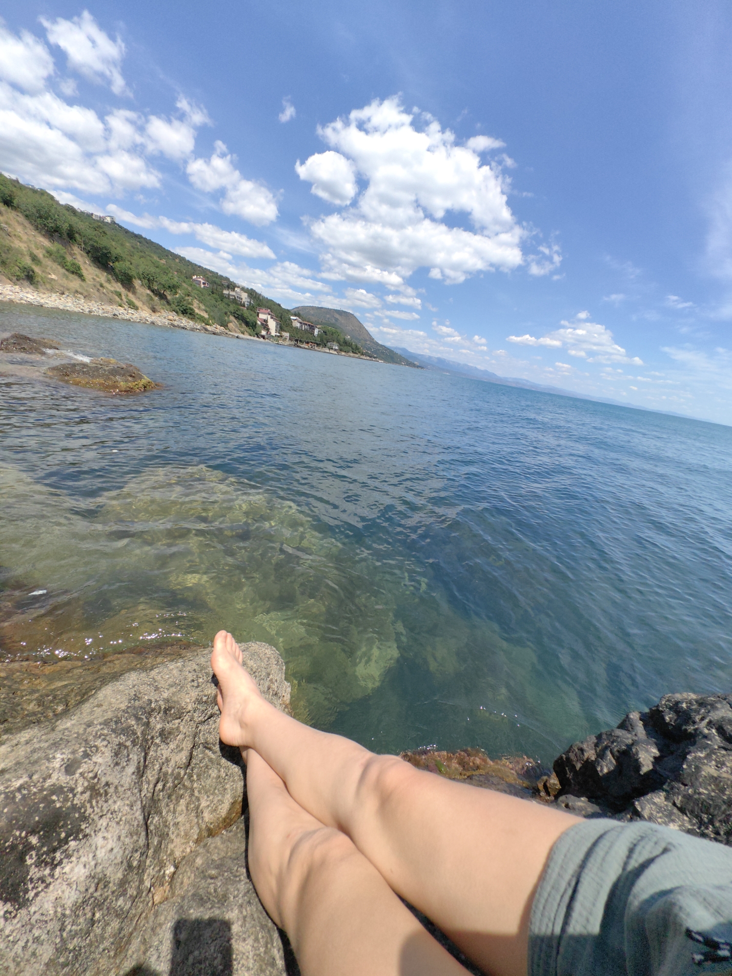 The image depicts a scenic view of the coast in Utios, Ukraine. In the foreground, a person's legs and feet are visible, crossed and resting on a large rock by the water's edge. The water is clear and blue, with some rocks visible beneath the surface. Beyond the water, a lush green hillside slopes down towards the sea, dotted with houses and trees. The sky above is a vibrant blue, adorned with fluffy white clouds, suggesting a pleasant, sunny day. The overall impression is one of tranquility and natural beauty.