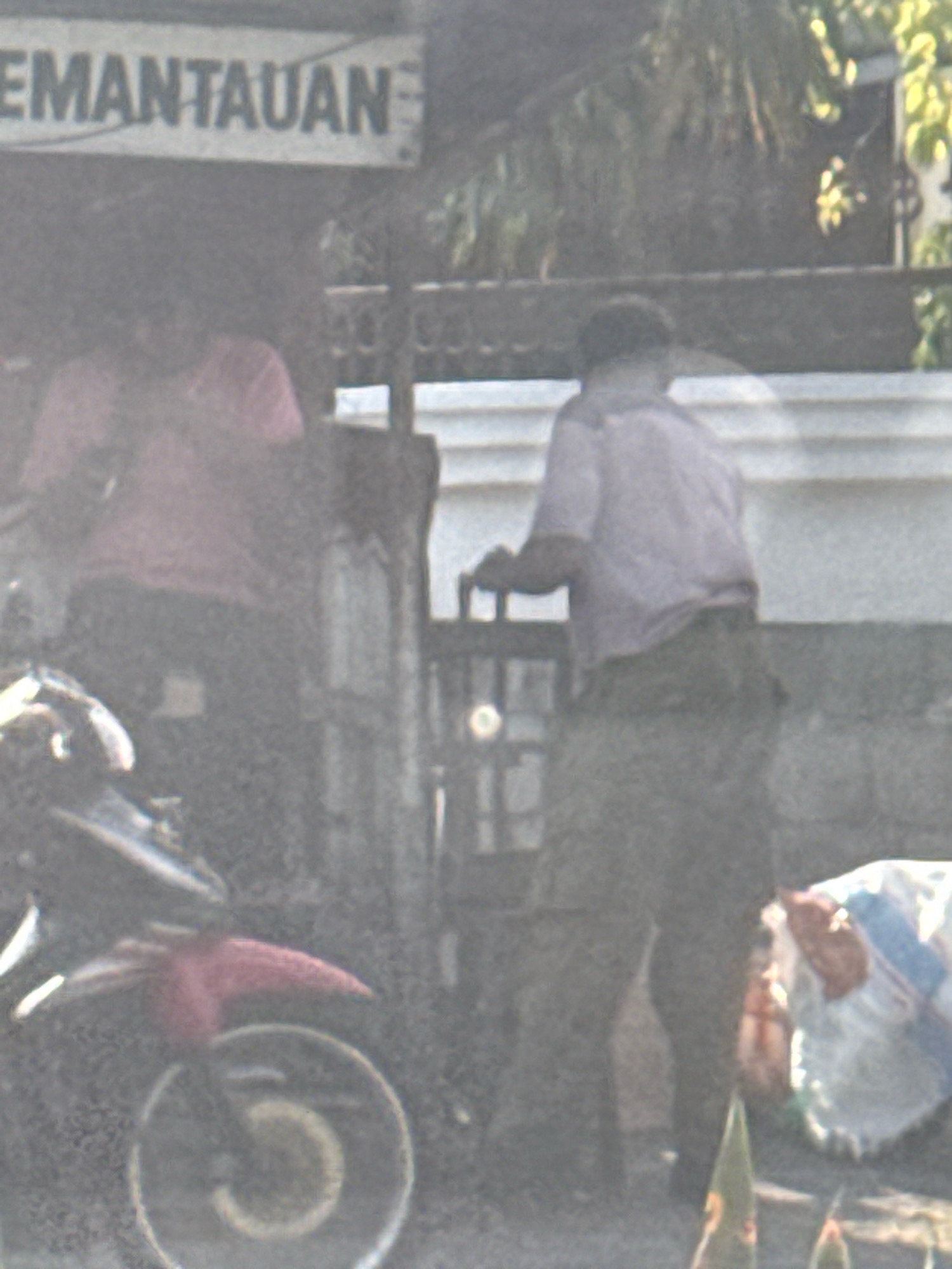 The image depicts an outdoor scene, likely a street or an area with a building. In the foreground, a motorcycle is partially visible, with its front wheel and a red fender in focus. Behind the motorcycle, two individuals are present. One person, wearing a light purple shirt and dark pants, is standing and appears to be interacting with a gate or a fence. Another person, wearing a pink shirt, is seated or partially visible in the background, behind the gate. A sign with the word "EMANTUAN" is visible at the top of the image, suggesting it might be a business or a service establishment. The overall image is slightly blurry and has a hazy quality. There are also some plants visible in the lower right corner of the frame. The scene appears to be located in Surabaya, Indonesia, as indicated by the context provided.