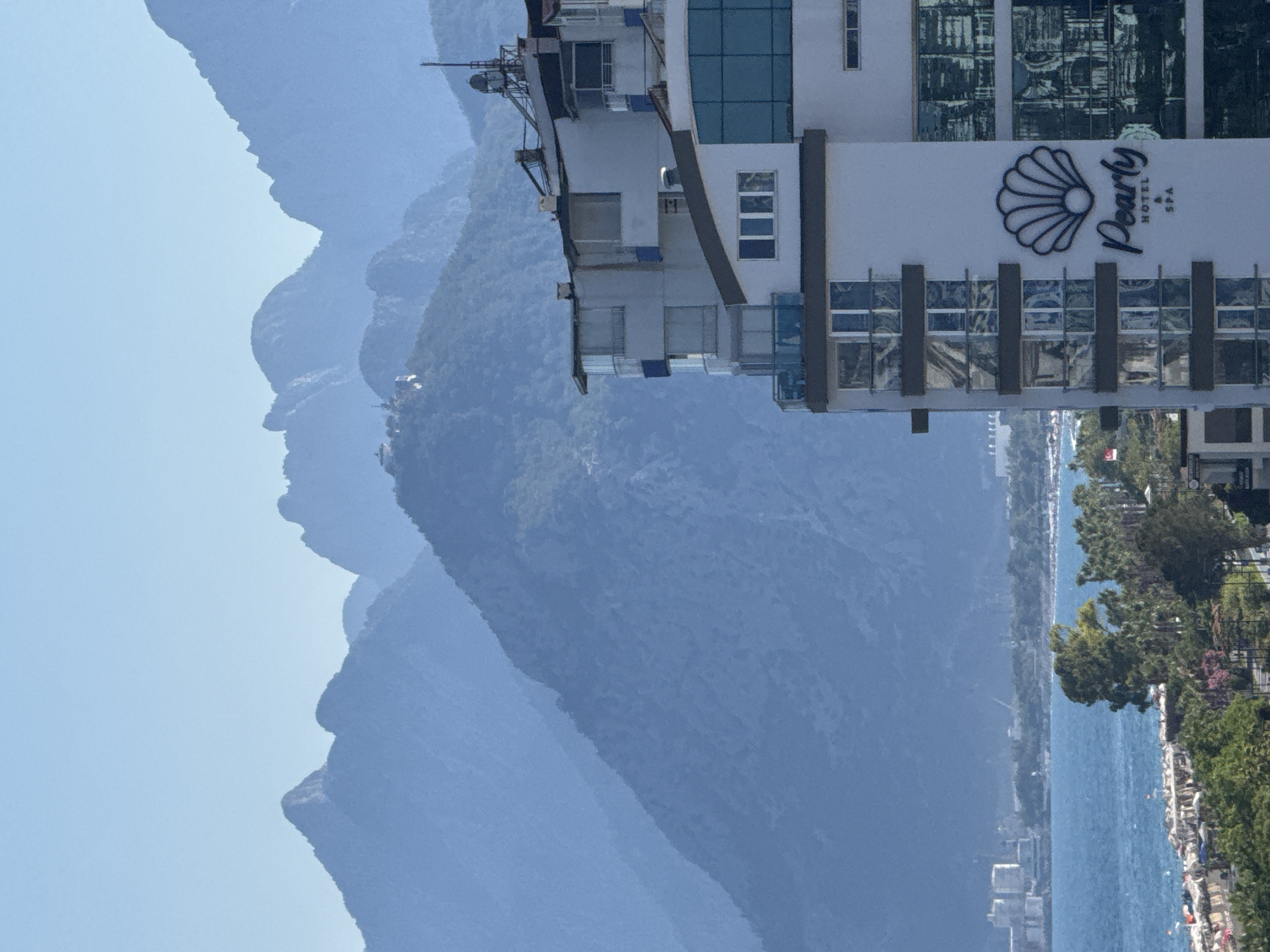 The image shows a hotel building with the text "Pearly HOTEL SPA" and a shell logo. The hotel is situated on a hill overlooking a body of water, likely the sea, with a shoreline lined with trees and buildings. In the background, a series of mountains rise, with the closest one having a prominent, face-like profile. The sky is a clear, light blue, suggesting fair weather and daytime. The scene appears to be a tourist or resort area. There are no people visible in the image, but there are some structures and natural elements that suggest human presence in the vicinity. The image is oriented vertically.