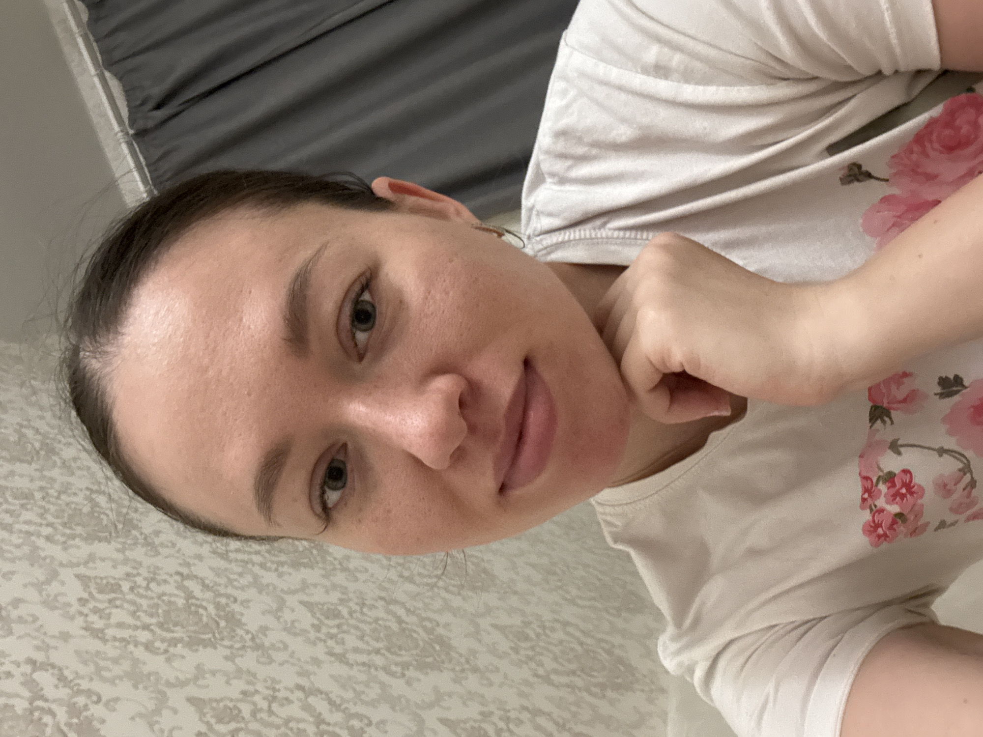 A woman is posing for a selfie indoors. She has fair skin, brown hair pulled back, and is wearing a white t-shirt with a floral design. Her right hand is touching her chin. The background consists of a wall with a patterned wallpaper and a grey curtain. The image was taken in Ферма, Russia.