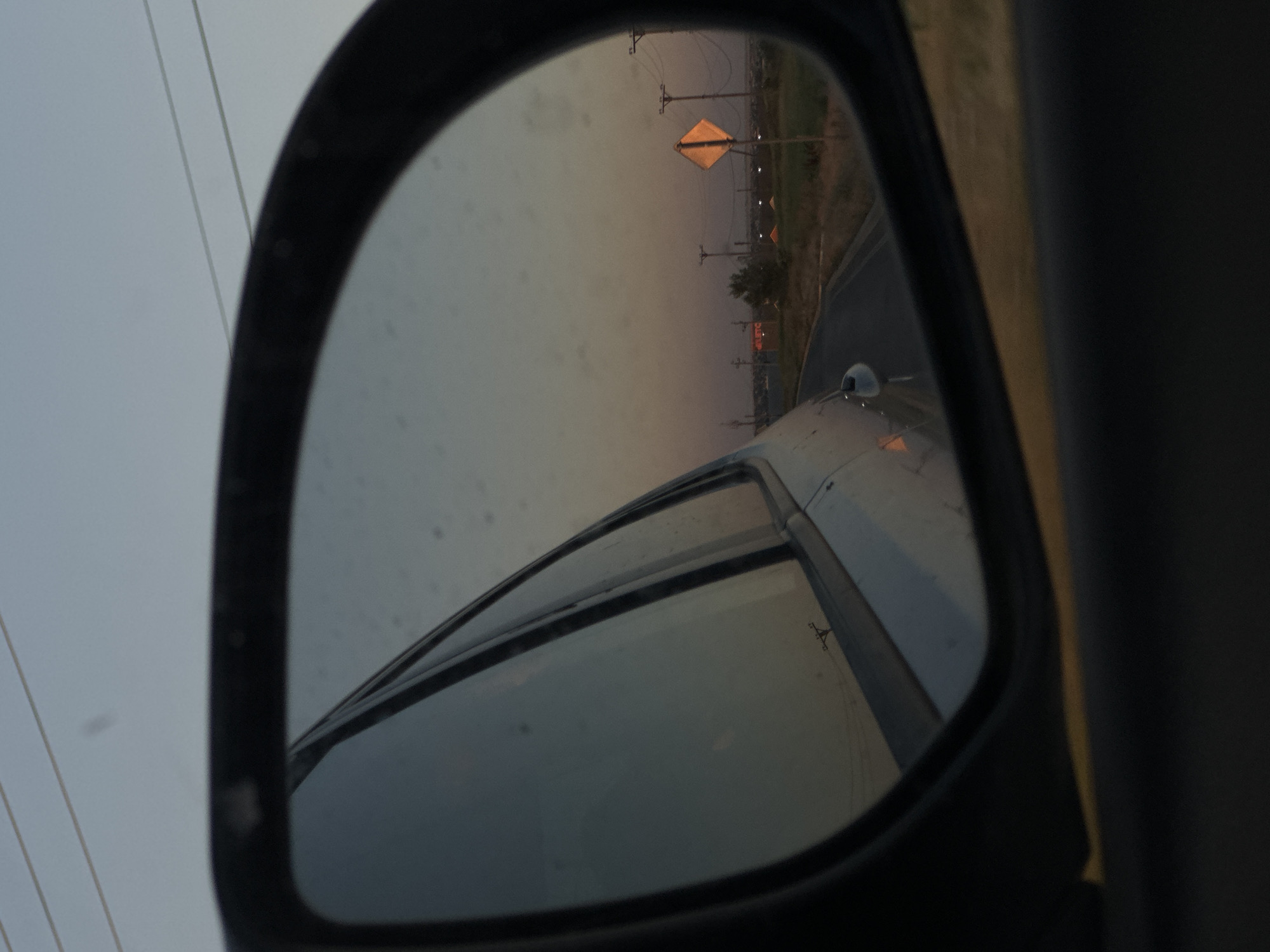 This media file shows a reflection in a car's side mirror. The reflection captures a road with a diamond-shaped orange sign, likely indicating a hazard or warning. Several utility poles with wires are visible along the roadside. In the distance, there appear to be buildings or structures, one of which has an orange hue, possibly indicating construction or a specific type of building. The sky is overcast, suggesting a cloudy or hazy day, and the light indicates it is either early morning or late afternoon, as the sun is low on the horizon. The scene is somewhat rural or suburban, with open land beside the road. The reflection in the mirror also shows the side of the car from which the picture is taken, including part of the vehicle's body and window. There is no visible text in the image.