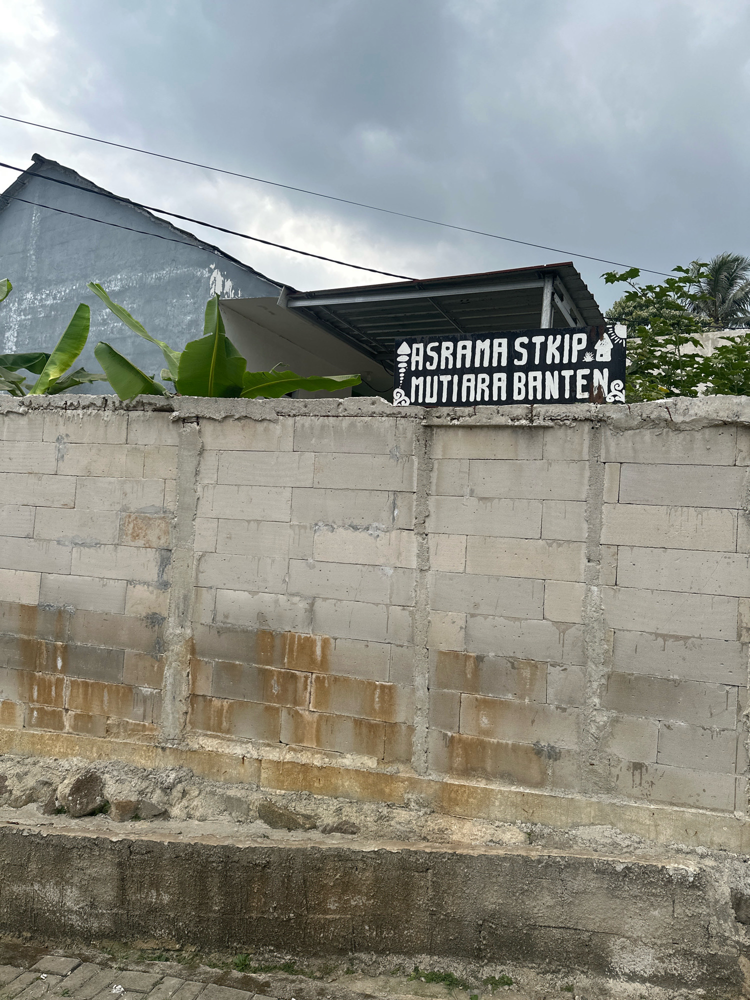 A black sign with white lettering reads "ASRAMA STKIP MUTIARA BANTEN" and is mounted on top of a weathered concrete block wall. The wall is segmented by vertical concrete supports and shows signs of water staining. Behind the wall, a gray house with a sloping roof and banana trees can be seen. Power lines crisscross the overcast sky. The scene is an outdoor environment in Saruni, Indonesia.