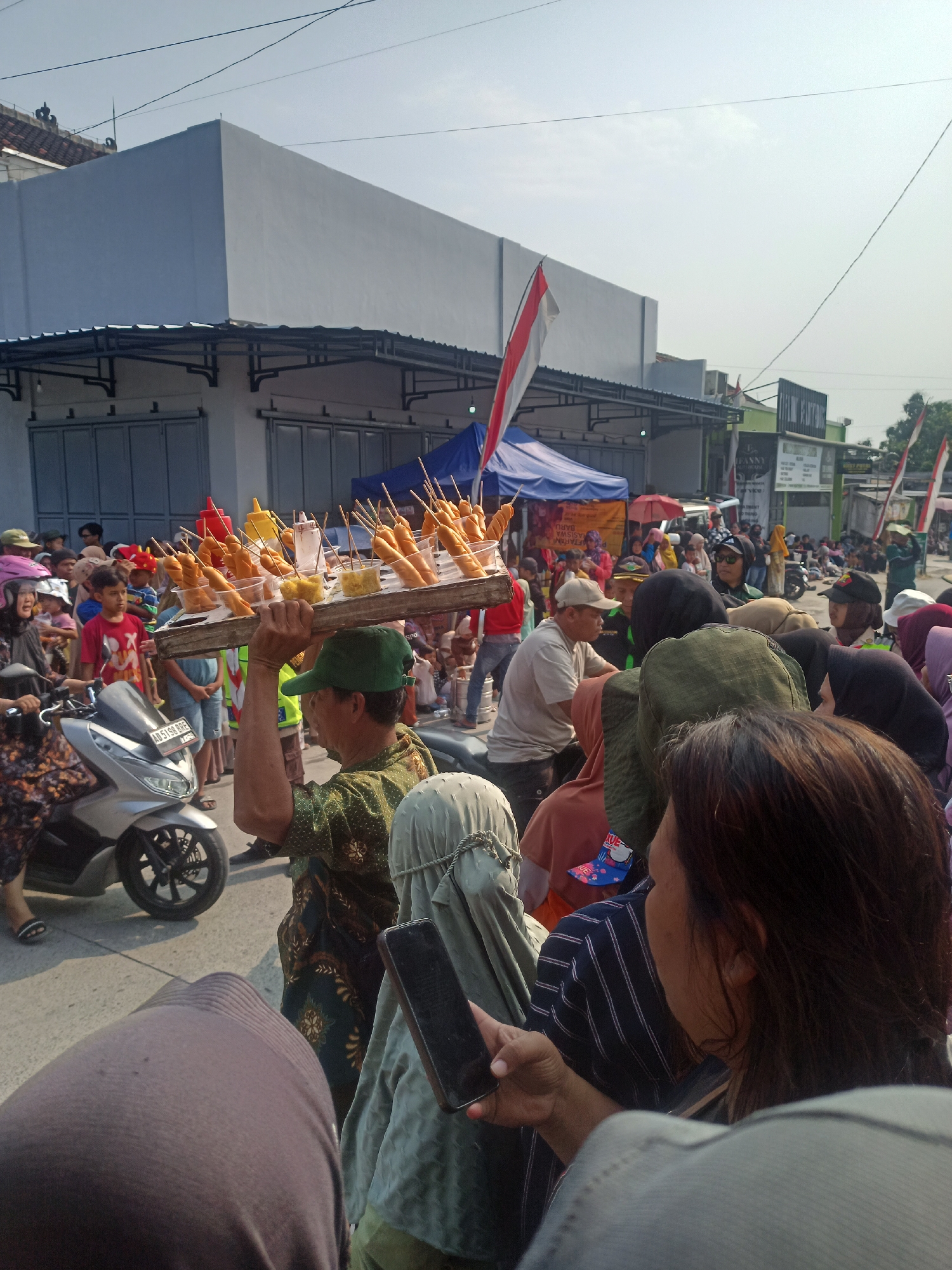 The image depicts a street scene in Gemolong, Indonesia, under a clear sky. In the foreground, a person with dark hair is holding a smartphone and looking to the right. Their shoulder and part of their head are visible, and they are wearing a striped shirt. To their left, another person wearing a brown headscarf and green hijab is partially visible. Directly in front of them, a person wearing a brown headscarf and a dark grey ribbed garment is seen from the back.
In the midground, a man wearing a green cap and a patterned shirt is carrying a wooden tray laden with fried snacks on skewers. These snacks appear to be sausages or similar items, presented in plastic cups with condiments. A silver scooter is parked to the left of the man. Behind him, a crowd of people are gathered, some in the shade of awnings.
The background features a long, light-colored building with a corrugated metal roof. Several Indonesian flags, red and white, are visible, suggesting a festive occasion or national celebration. Tents and market stalls are also present, indicating a public gathering or event. The street is paved, and there are power lines overhead. The overall atmosphere appears to be that of a busy outdoor market or festival.