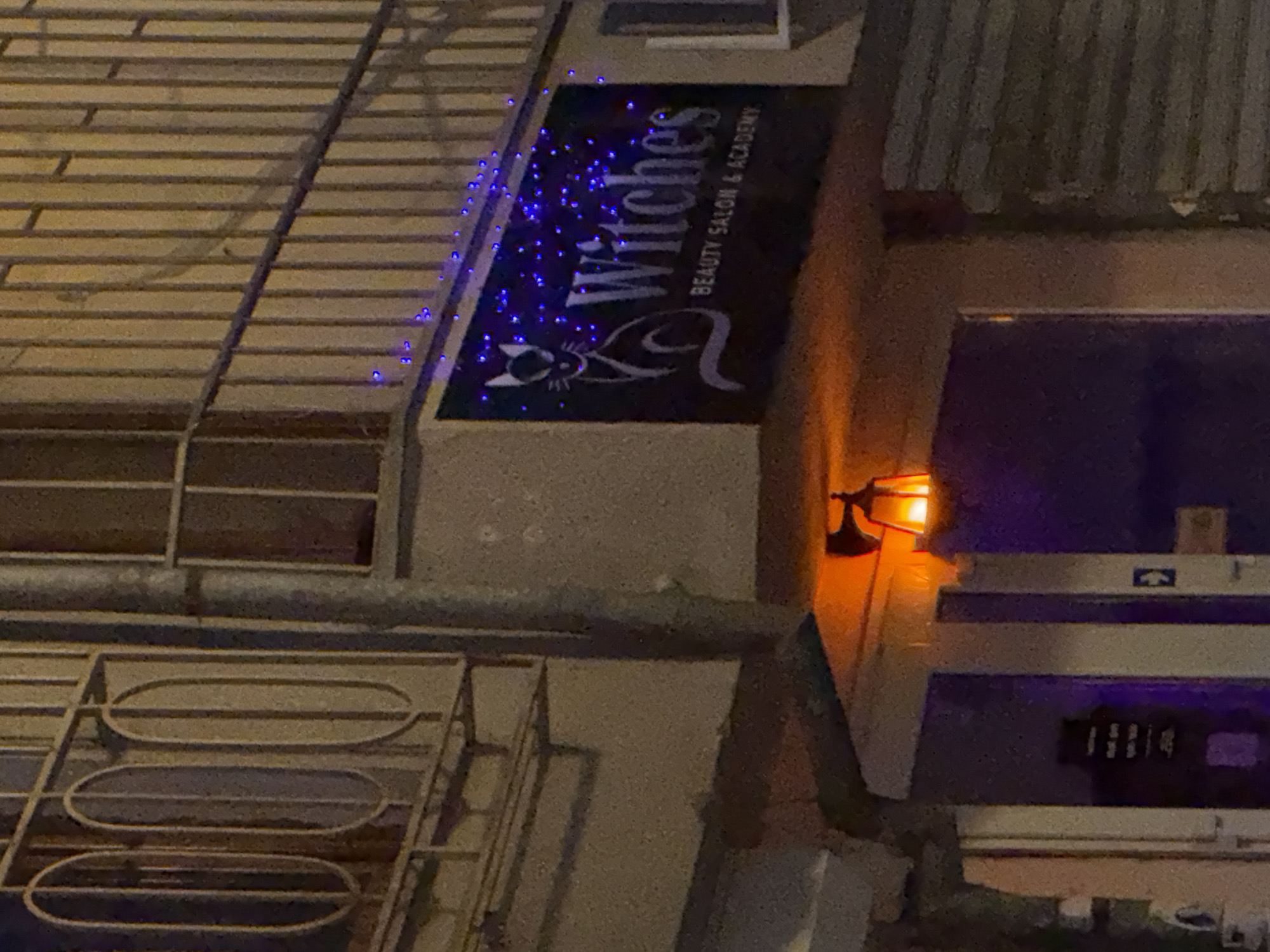 The image shows the exterior of a building, likely located in Varna, Bulgaria. The building has a sign that reads "Witches, Beauty Salon & Academy." The sign is decorated with blue lights. A black cat is incorporated into the logo on the sign. There is a light fixture near the sign, casting an orange glow. The building appears to have bars on the windows and a metallic exterior. A darker window is visible on the side of the building.