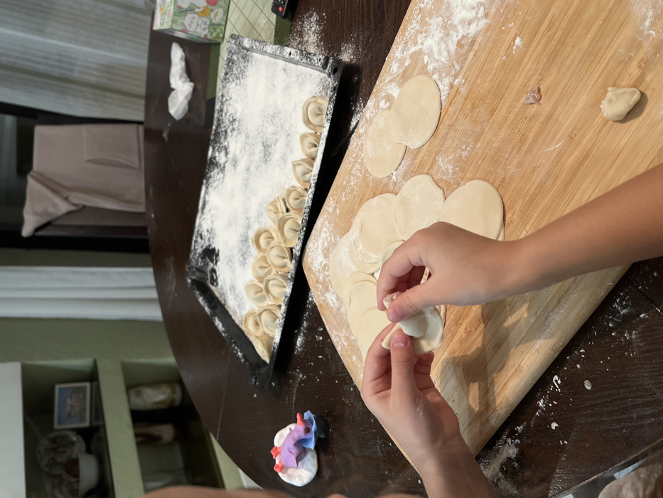 The image depicts a domestic scene in what appears to be a kitchen or dining area within a residence in Krasnoyarsk, Russia, centered on the preparation of pelmeni. A pair of human hands, likely belonging to a child based on size, occupies the lower-left foreground, actively folding a circular piece of dough to form a dumpling. A light-colored wooden cutting board, dusted with white flour, holds multiple flattened, circular dough wrappers to the right of the hands, along with a small, completed pelmen and a tiny piece of raw meat filling.
To the upper right of the cutting board, a black rectangular baking tray, heavily coated with flour, contains a substantial quantity of already-formed pelmeni arranged in rows and clusters. The tray rests on a dark, polished wooden table, which is also lightly dusted with flour and bears some scattered crumbs.
In the mid-ground, to the left of the baking tray, a small, multi-colored figurine, possibly a toy, is visible on the table. Further into the background, on the right side of the table, a remote control, a small patterned box, and a crumpled white tissue are present. A light-colored towel or robe is draped over the back of a chair in the distant background. A light green wall with a built-in shelf unit is visible on the far left, displaying decorative plates, a small framed picture, and other ceramic items.