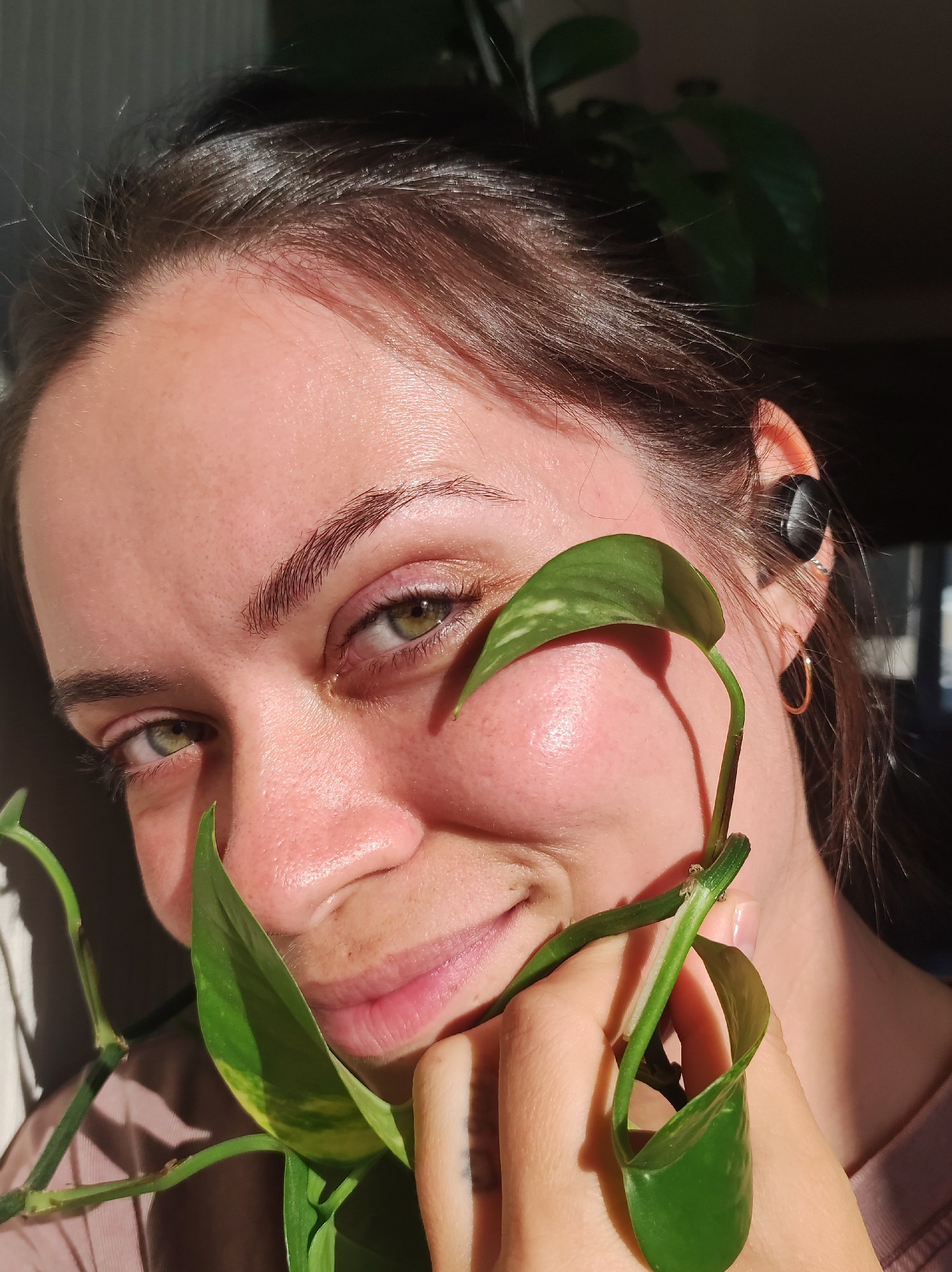 A close-up portrait shows a woman engaging directly with the camera, illuminated by strong, directional sunlight. She has dark brown hair, pulled back, and captivating green eyes. A gentle smile graces her lips, and her gaze is steady. The light from the right casts bright highlights across her forehead, cheeks, and the bridge of her nose, making her skin appear glistening or dewy.
She is holding a vine of a pothos plant, characterized by its glossy, heart-shaped green leaves, some displaying yellow variegation. One significant leaf is positioned near her mouth, and the vine and other leaves cast distinct shadows on her face and shoulder. In her right ear, a black wireless earbud is visible, accompanied by a small gold hoop earring in her earlobe. Her eyebrows are neatly shaped and dark.
The background is dark and blurred, suggesting a shallow depth of field, bringing the woman and the plant into sharp focus. A light-colored garment, possibly a t-shirt, is visible on her shoulder and around her neck. The harsh, direct sunlight indicates a daytime setting, likely indoors near a window. There is no visible text in the image. The overall impression is one of natural beauty and serenity.