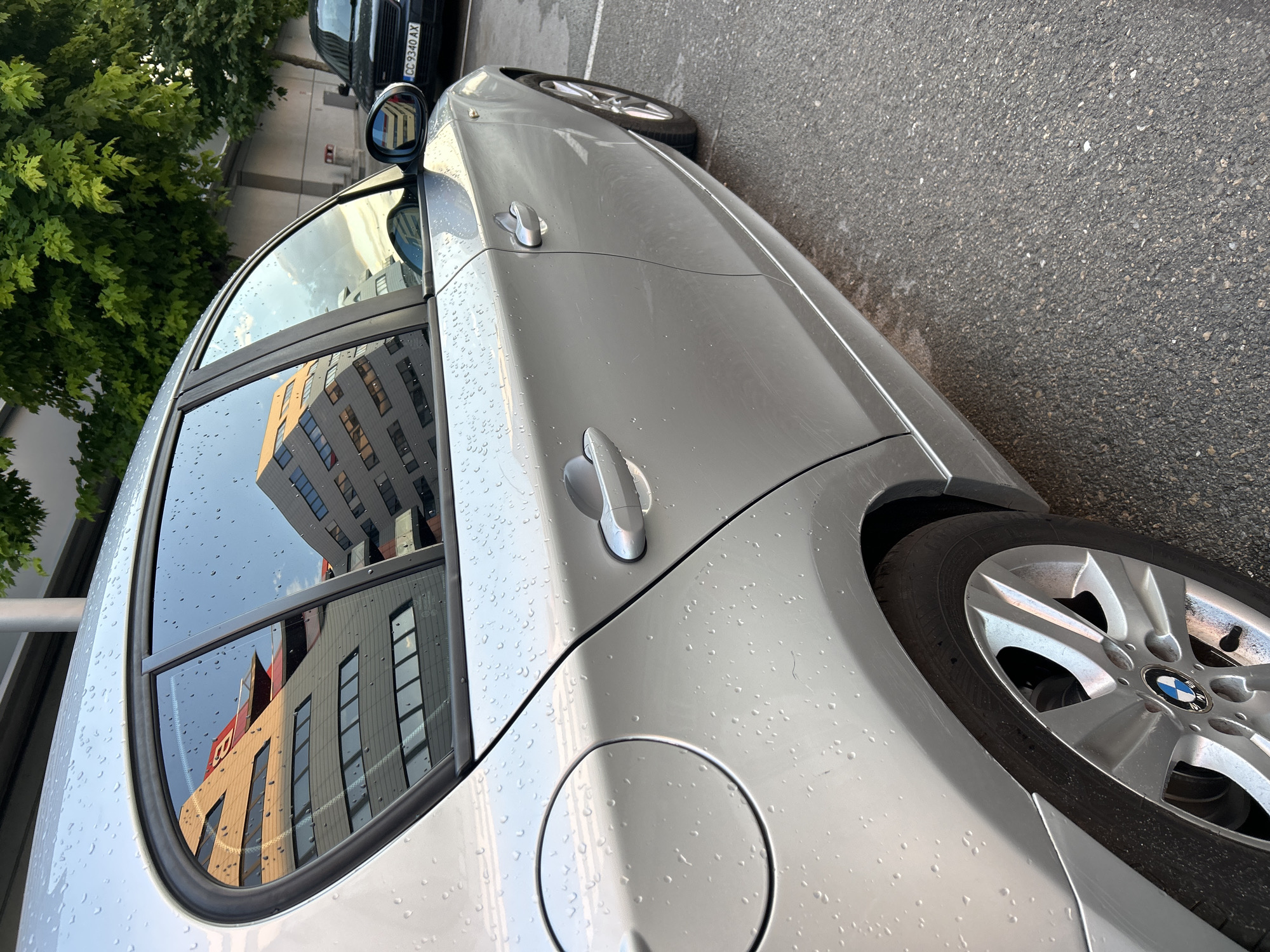 The image shows a side view of a silver BMW parked on an asphalt surface. The car's body is wet with raindrops, and reflections of a modern building with red accents are visible on the windows. In the background, a dark-colored car with a Bulgarian license plate "CC 9340 AX" is parked, and green trees are present. The scene appears to be an outdoor parking lot, possibly in the city of Ruse, Bulgaria, given the license plate. There are no people visible in the image.