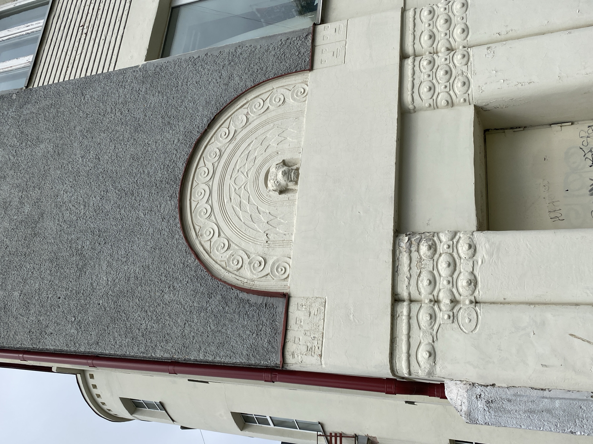 The image shows a close-up, exterior, eye-level view of a light-colored building facade with decorative elements in Sortavala, Russia. The building has a rough grey textured upper section and a smoother, lighter colored section below. A prominent feature is a semi-circular decorative relief above a doorway. This relief contains a central grotesque face, possibly a lion or dog, surrounded by concentric circular patterns and geometric designs. To the left of the grey section is a curved tower-like structure with small, square-topped windows. A dark red pipe runs vertically along the side of the building. The lower portion of the facade features a doorway and decorative pilasters with repeating oval and circular motifs. The building appears aged, with peeling paint visible on some surfaces. The overall architectural style suggests an Art Nouveau or similar early 20th-century influence.