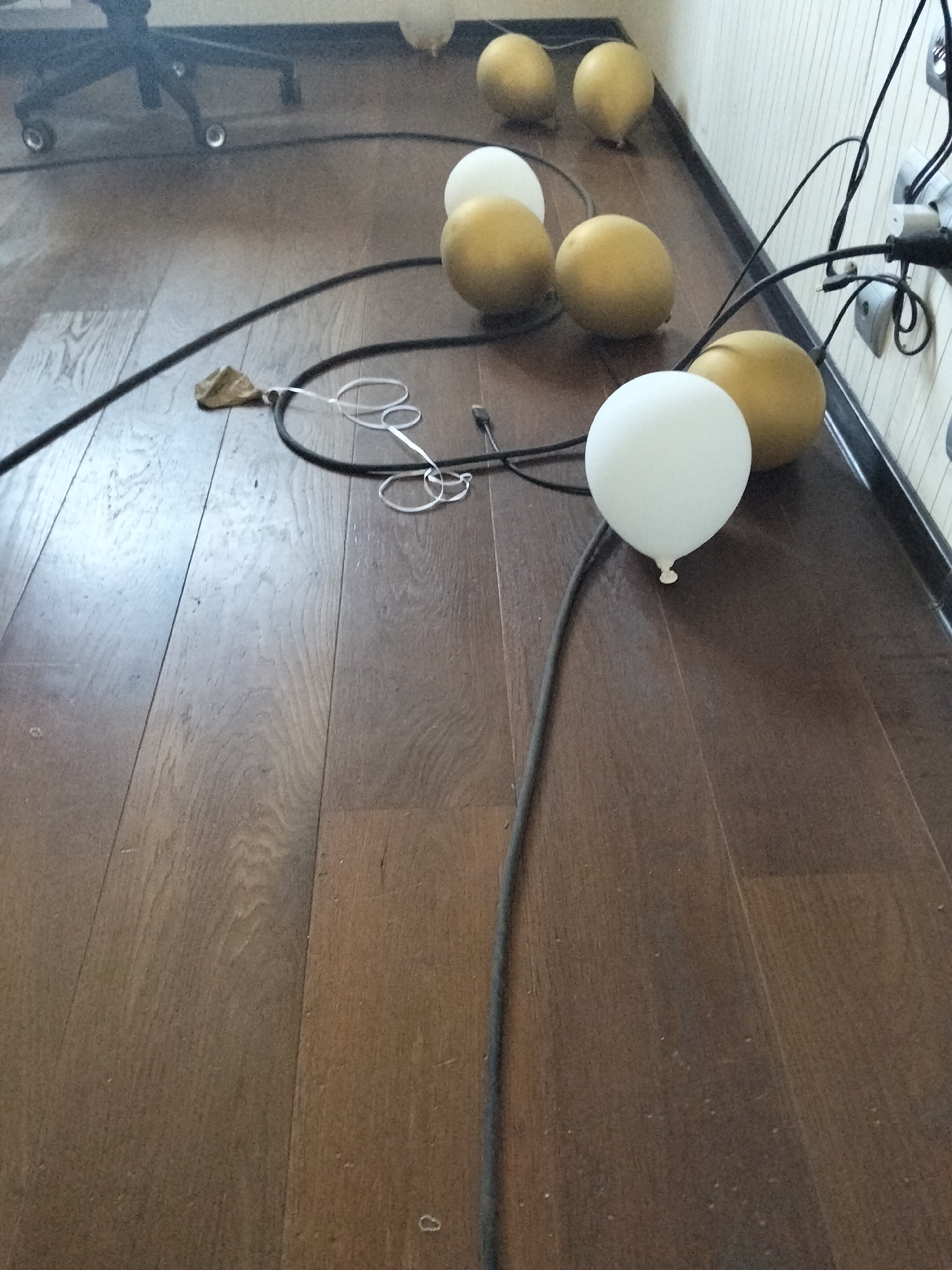 The image shows a floor with several balloons scattered on it. There are also black cables and a power strip with multiple outlets visible. The floor appears to be made of wooden planks. The balloons are a mix of white and gold. Some of the balloons are connected to the cables. There is a black office chair with wheels in the background, slightly out of focus. The scene is indoors and the lighting suggests it is daytime.