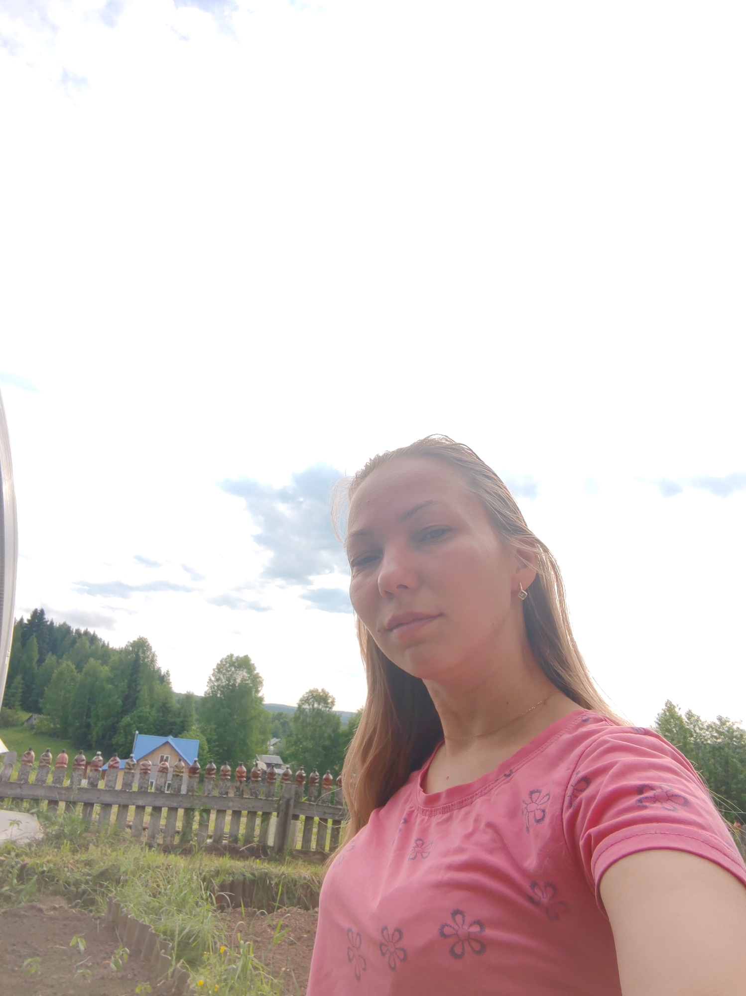 A young woman with long, blonde hair is taking a selfie outdoors. She is wearing a pink t-shirt with a floral pattern and a thin gold necklace. Her face is turned towards the camera, with a neutral expression.
In the background, there is a wooden fence with decorative elements on top. Behind the fence, a village scene unfolds, with houses and trees visible on a gentle slope. The sky is bright and overcast with some clouds. The overall impression is a casual, rural setting, likely in the city of Chuchlem, Russia, given the architectural style of the houses and the natural landscape.