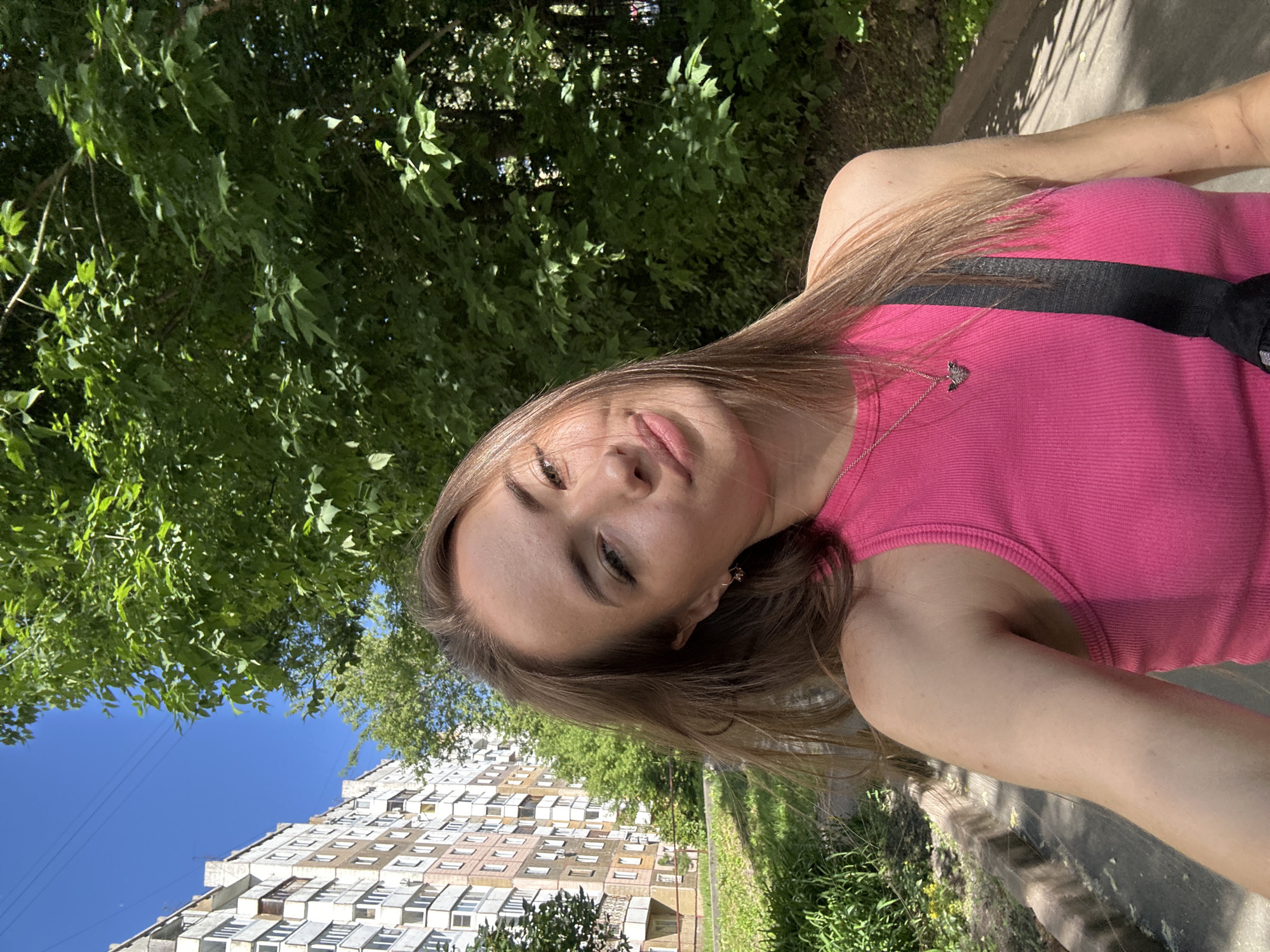 A young woman with long, light brown hair takes a selfie outdoors on a sunny day. She is wearing a bright pink ribbed tank top with a simple silver necklace featuring a butterfly pendant. A black strap, likely from a bag, is visible across her chest. Her left arm and shoulder are visible, showing some faint freckles.
In the background, a tall, multi-story apartment building with numerous windows and balconies is visible on the left. Green trees and lush foliage dominate the right side of the frame and extend across the upper background, creating a dappled sunlight effect on the pavement. The ground in the foreground appears to be a paved pathway or sidewalk with a curb. The sky is clear and blue. The overall scene suggests a casual outdoor setting within a residential urban area.