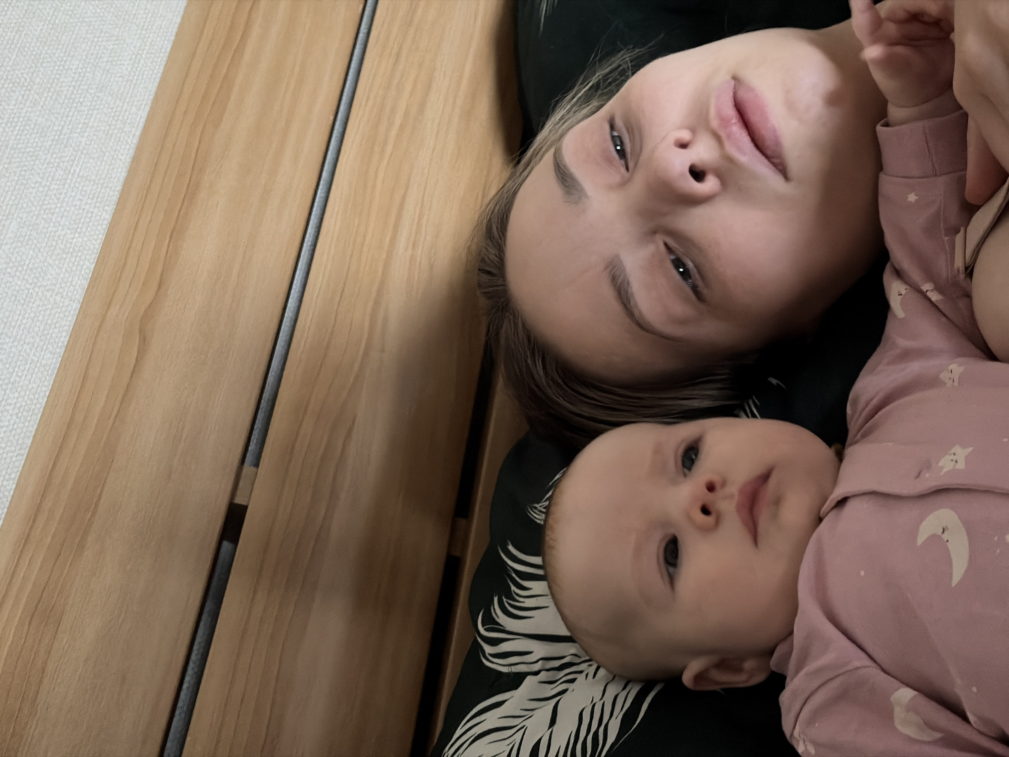An indoor, overhead shot captures an adult woman and an infant lying close together, likely on a bed or a comfortable surface. The image is oriented so the subjects' heads are towards the upper right.
The adult, a fair-skinned woman with dark hair, lies on her back, looking directly at the camera with a neutral to relaxed expression. Her eyes are open, and she has visible eyebrows. She appears to be resting on a dark pillow or sheet, which is also part of her clothing.
Below and slightly to the left of the woman, a fair-skinned infant lies on its back, looking upwards and slightly to the right with a calm, curious, or contemplative gaze. The baby is dressed in a light pink long-sleeved outfit featuring white and light grey celestial patterns, including moons and stars. Part of the woman's arm is visible near the baby's head, suggesting she is holding or comforting the child.
To the left of the subjects, a significant portion of the image is occupied by a light wooden surface, which could be part of a bed frame, wooden floorboards, or a piece of furniture. A dark, thin vertical groove runs through this wooden surface. Further to the far left, a textured white surface, possibly a wall or carpet, is visible.
The lighting is soft and appears to be artificial or from a dimly lit environment, suggesting the time of day could be evening or nighttime. There is no visible text in the image. The scene depicts a quiet, intimate moment in a domestic setting, characteristic of a home in locations such as Chaikovsky, Russia.