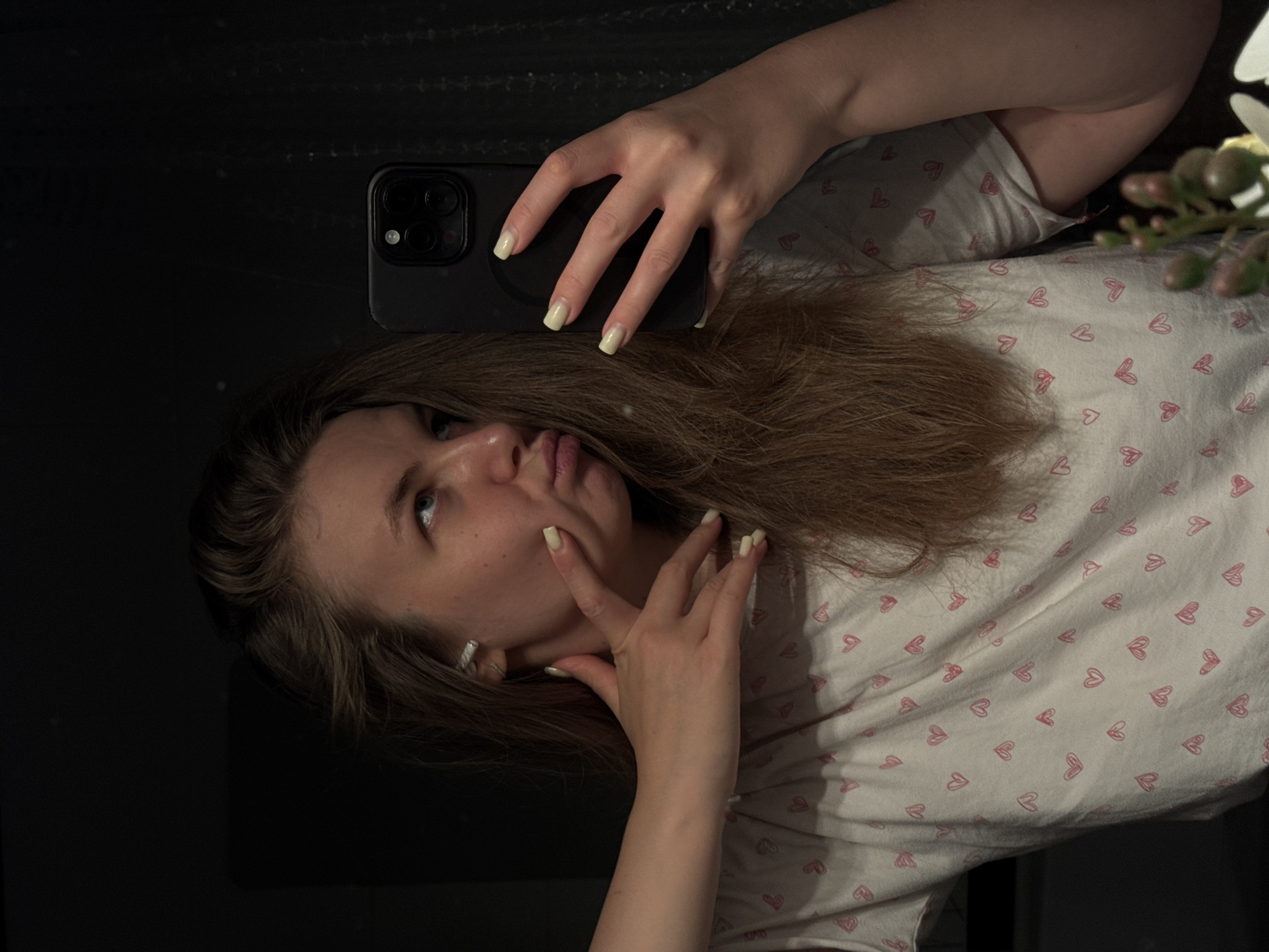 A young woman is holding a black smartphone up to take a selfie. She is positioned in front of what appears to be a mirror or a dark, reflective surface. She has light brown hair and is wearing a white t-shirt with small pink hearts. The camera of the phone is visible, as are her hands, which are holding the phone. Her fingernails are painted a light yellow color. The background is mostly dark, with a few decorative items visible in the upper right corner. There is no indication that this image was taken in Vladimir, Russia.