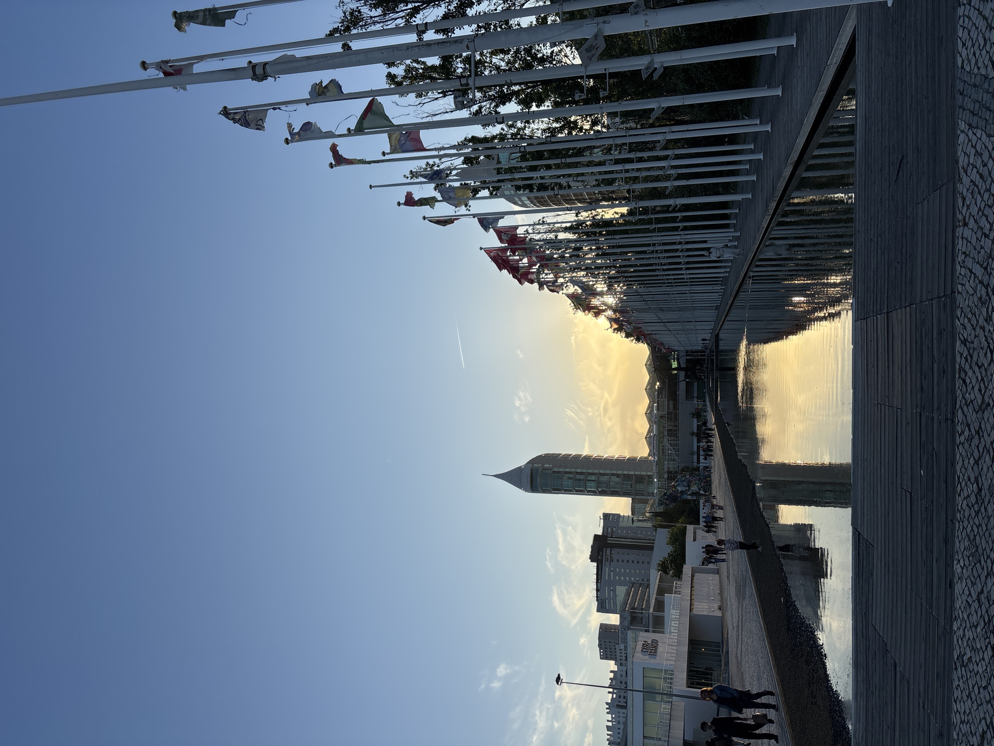 The image depicts a waterfront scene in Lisbon, Portugal, likely within or near the Parque das Nações area. A row of white flagpoles with tattered flags lines the upper portion of the frame. Below the flagpoles, a body of water reflects the sunset or early evening light. In the distance, a modern building with a distinctive spire is visible, as are other contemporary buildings. A walkway or promenade runs alongside the water, and people are walking. The lighting suggests either sunset or sunrise.