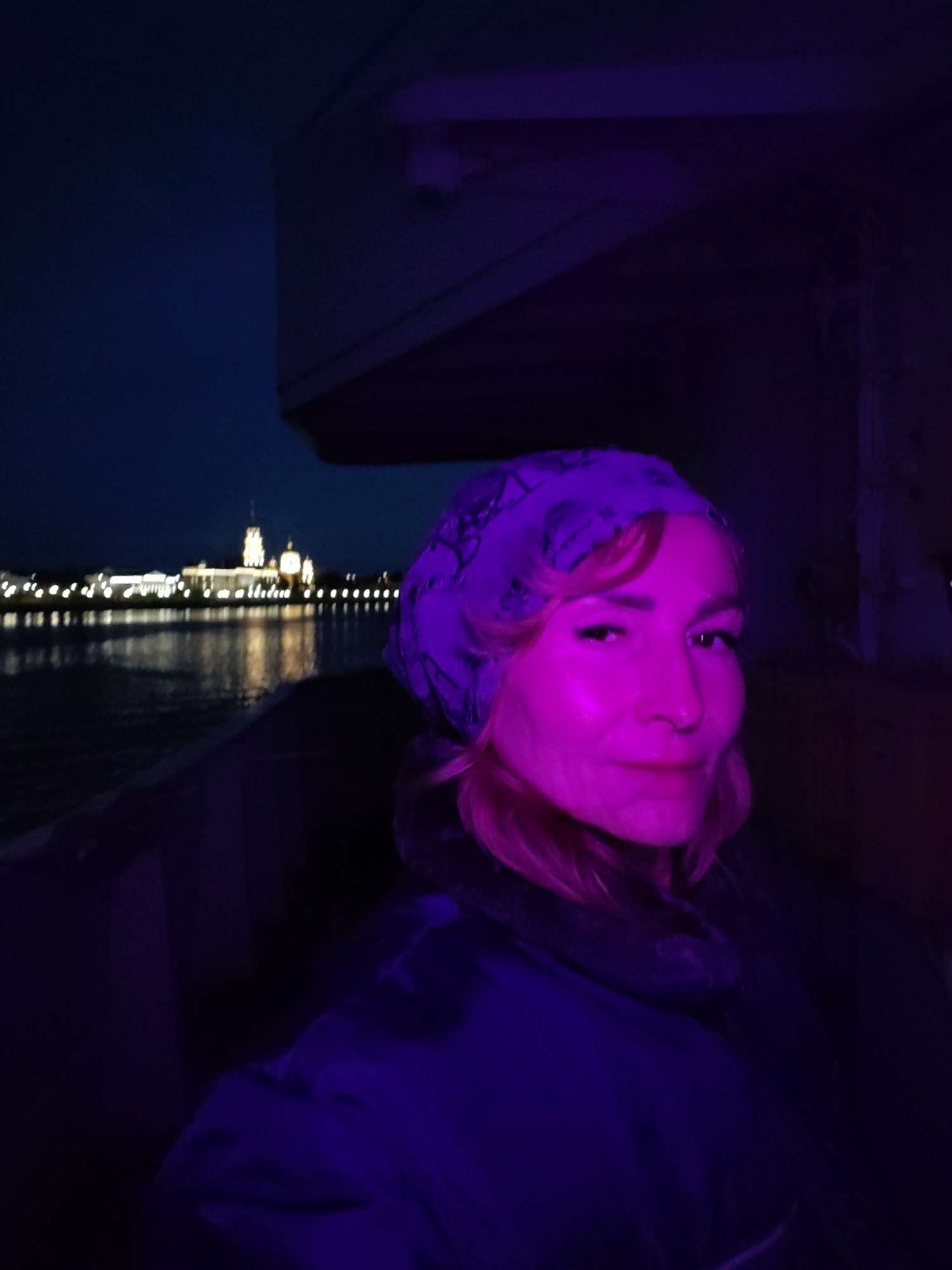 A woman is photographed at night, with her face illuminated by purple light. She is wearing a headscarf with a pattern and a dark jacket. In the background, a city skyline with illuminated buildings is visible across a body of water. The overall scene suggests an outdoor evening setting, possibly on a boat or near a waterfront. The location is identified as Bol'shie Mkhi, Russia.