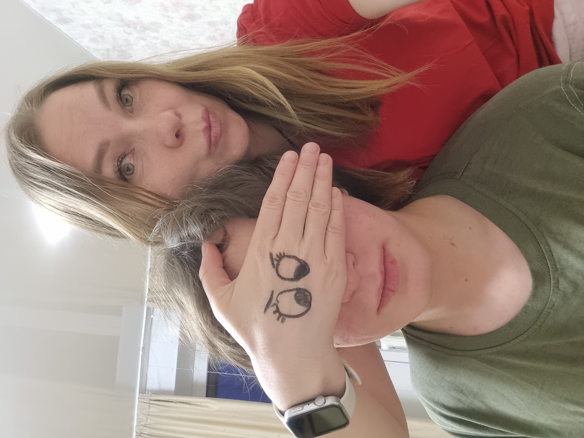 Two Caucasian women are in an indoor setting. One woman, with blonde hair and a red shirt, is in the foreground looking at the camera. The other woman, wearing a green shirt, has her eyes covered with her hand. Two drawn eyes are on the back of the hand. A white smartwatch is on the wrist of the hand that covers the eyes. The location is Chaikovsky, Russia.