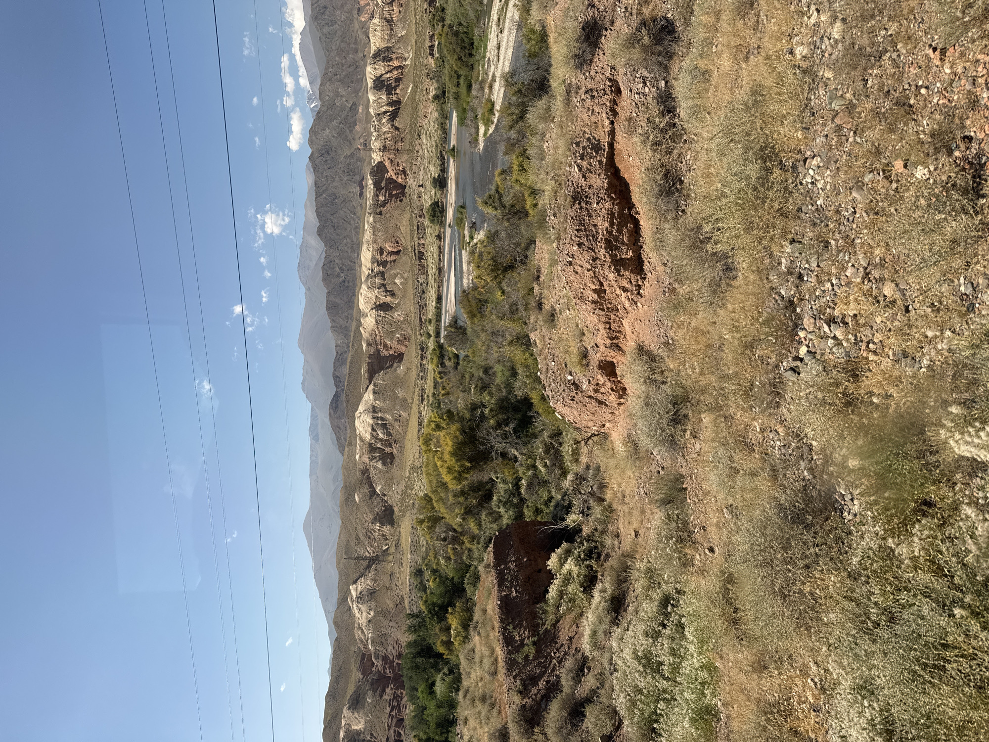 The image depicts a panoramic view of a mountainous and arid landscape. In the foreground, dry, scrubby vegetation and reddish-brown earth dominate the terrain. A small, winding river or stream flows through the middle ground, flanked by sparse green trees and shrubs. The background features layered mountains with exposed rock formations, exhibiting shades of tan, white, and red, indicative of erosion and geological strata. The sky is a clear, pale blue with a few wispy white clouds. Several thin, dark electrical wires stretch horizontally across the upper portion of the image, suggesting the presence of human infrastructure in the vicinity. The overall impression is one of a remote, natural environment under a bright, sunny sky. The location is identified as Unknown, Kyrgyzstan.