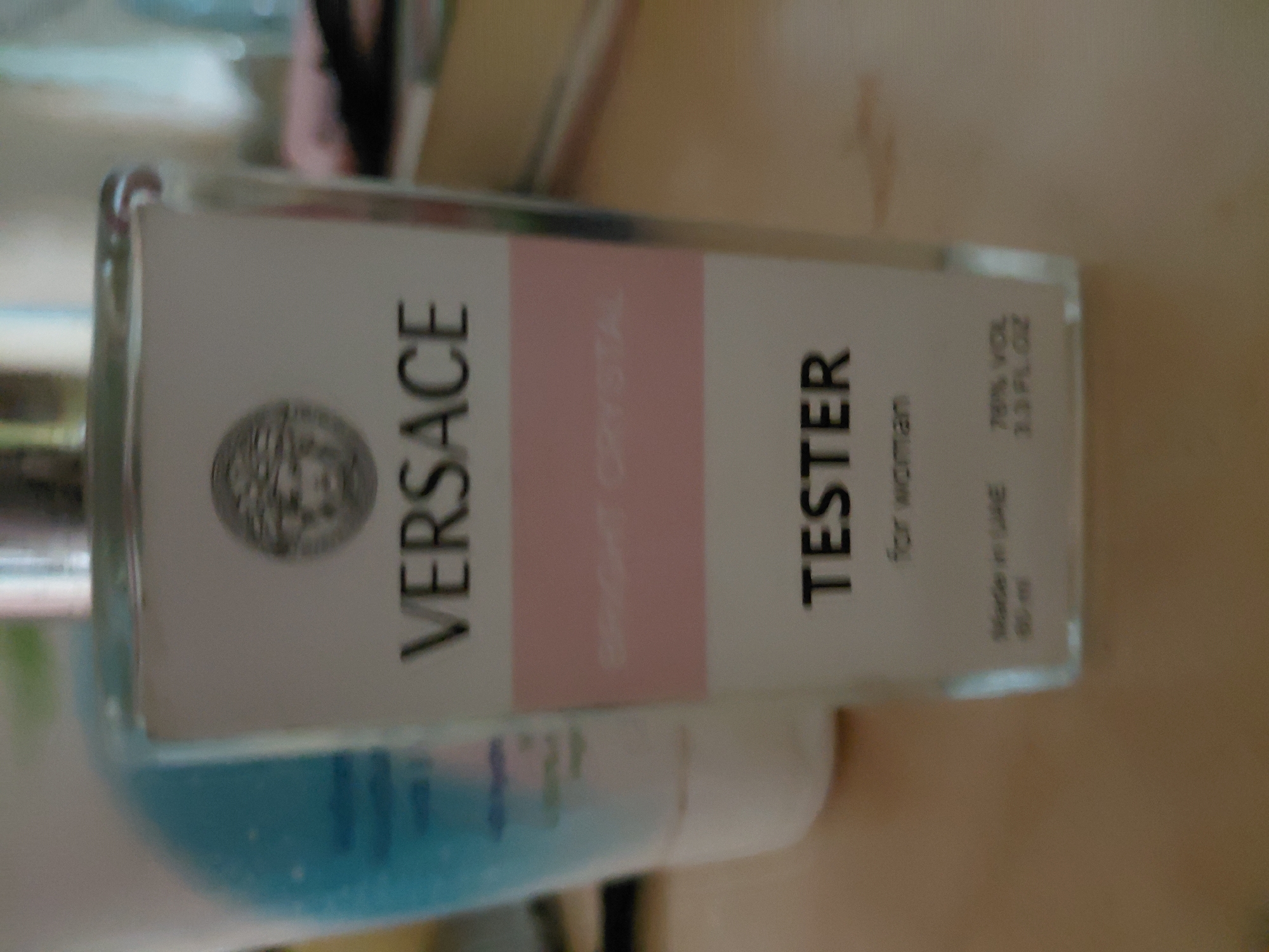The image is a close-up, slightly blurry, indoor shot of a Versace Bright Crystal Tester perfume bottle. The bottle is predominantly white with a pink stripe across the middle. The Versace logo, a Medusa head, is visible at the top of the label. Below the pink stripe, the word "TESTER" is written in large, black letters, followed by "for woman" in smaller text. At the bottom of the bottle, there are inscriptions that read "Made in UAE" and "70% VOL 3.3 FL.OZ.". The bottle is placed on a light-colored surface, likely a table. The background is out of focus, showing hints of other objects, possibly other cosmetic items or household items. The overall lighting is soft and diffused. There are no people visible in the image. The location is stated as Marynivka, Ukraine, but this information is not visually evident from the image itself.
