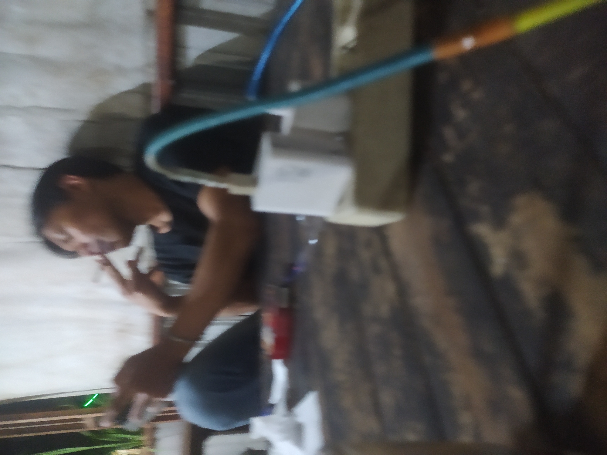 In the city of Tuntang, Indonesia, a person is seated at a wooden table, holding a cigarette and a mobile phone. The person is wearing a dark t-shirt and appears to be exhaling smoke from the cigarette. On the table, a red cigarette pack is visible, along with a white electrical adapter plugged into a power strip. A blue cable extends from the power strip, and the overall lighting suggests an indoor or dimly lit outdoor setting. The background shows a white wall with vertical wooden elements, and some green foliage is visible on the left side.