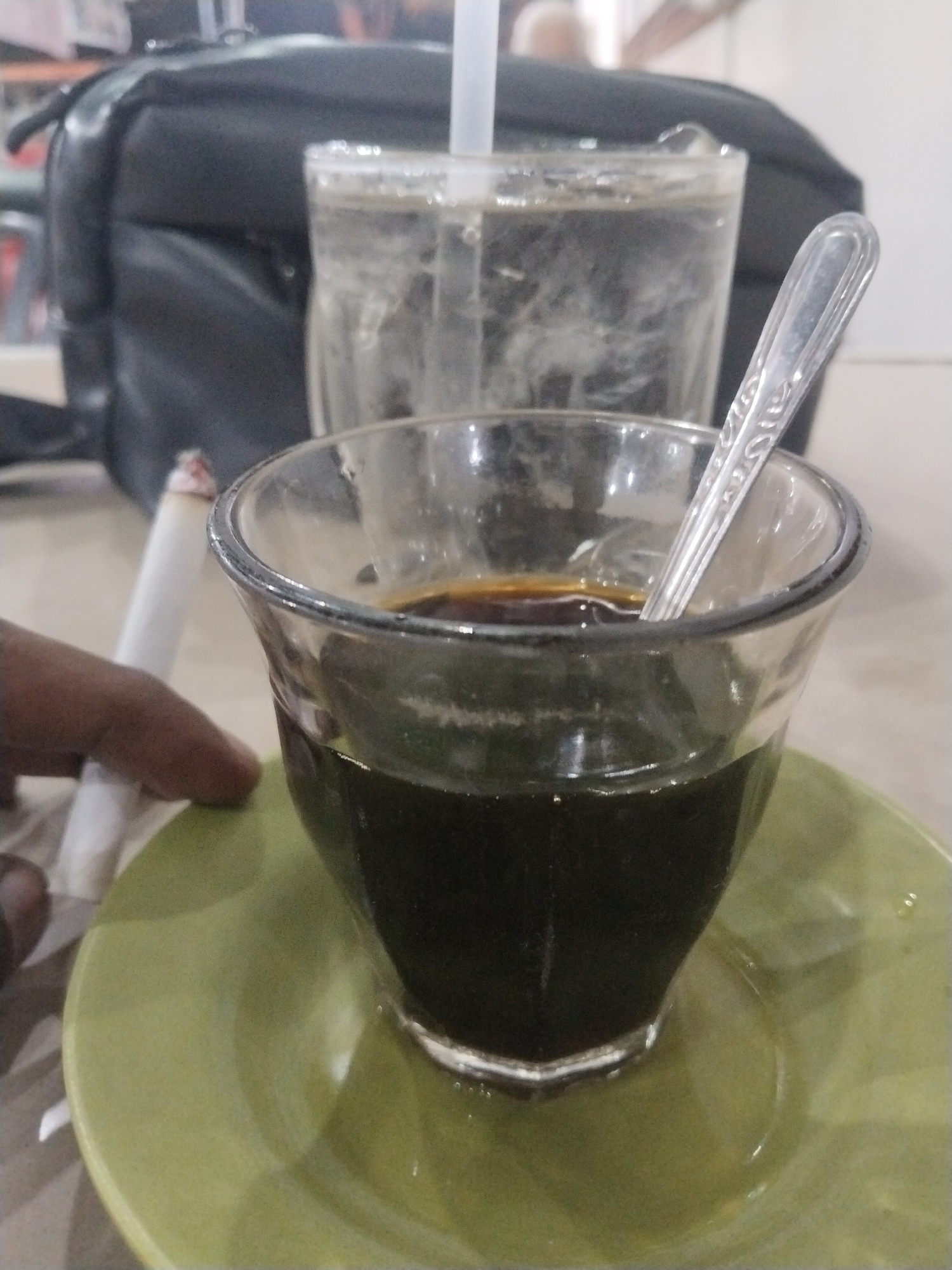 The image shows a table setting with a cup of coffee, a glass of water with a straw, and a lit cigarette. A hand is visible near the cigarette. A black bag is in the background. The setting is likely in Lhokseumawe, Indonesia, given the request context.