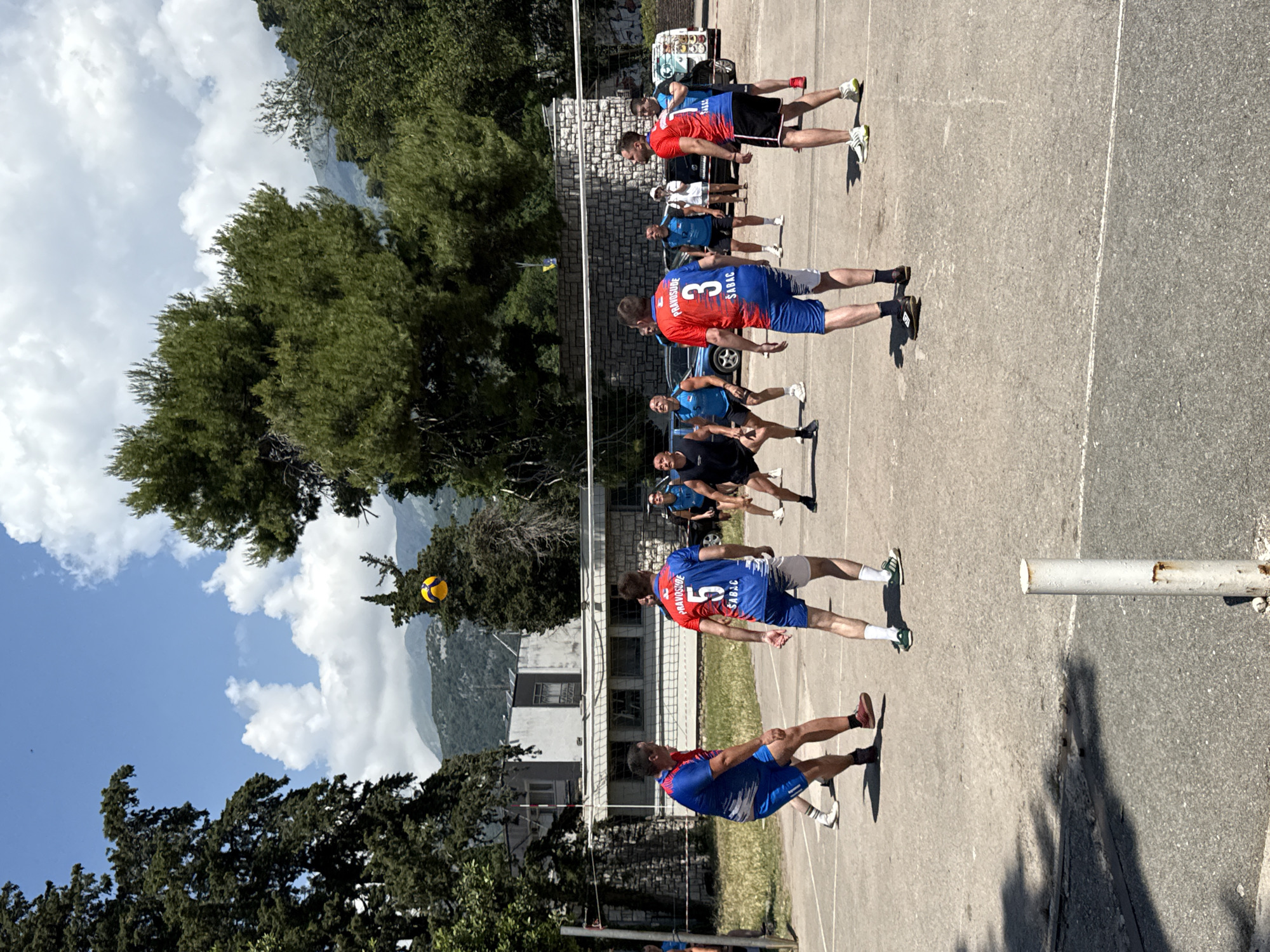 In Boreti, Montenegro, on a sunny day, a group of men are playing volleyball on an outdoor court. The court appears to be made of concrete with white lines marking its boundaries. A net divides the court in half, and players are positioned on both sides, actively engaged in the game. The volleyball is in the air, high above the net, indicating an ongoing play. The players are wearing athletic attire, with some in blue and red jerseys and shorts, and others in darker colored shorts and tops. The background features lush green trees, a stone wall, and buildings, with mountains visible in the distance under a bright blue sky with scattered clouds.