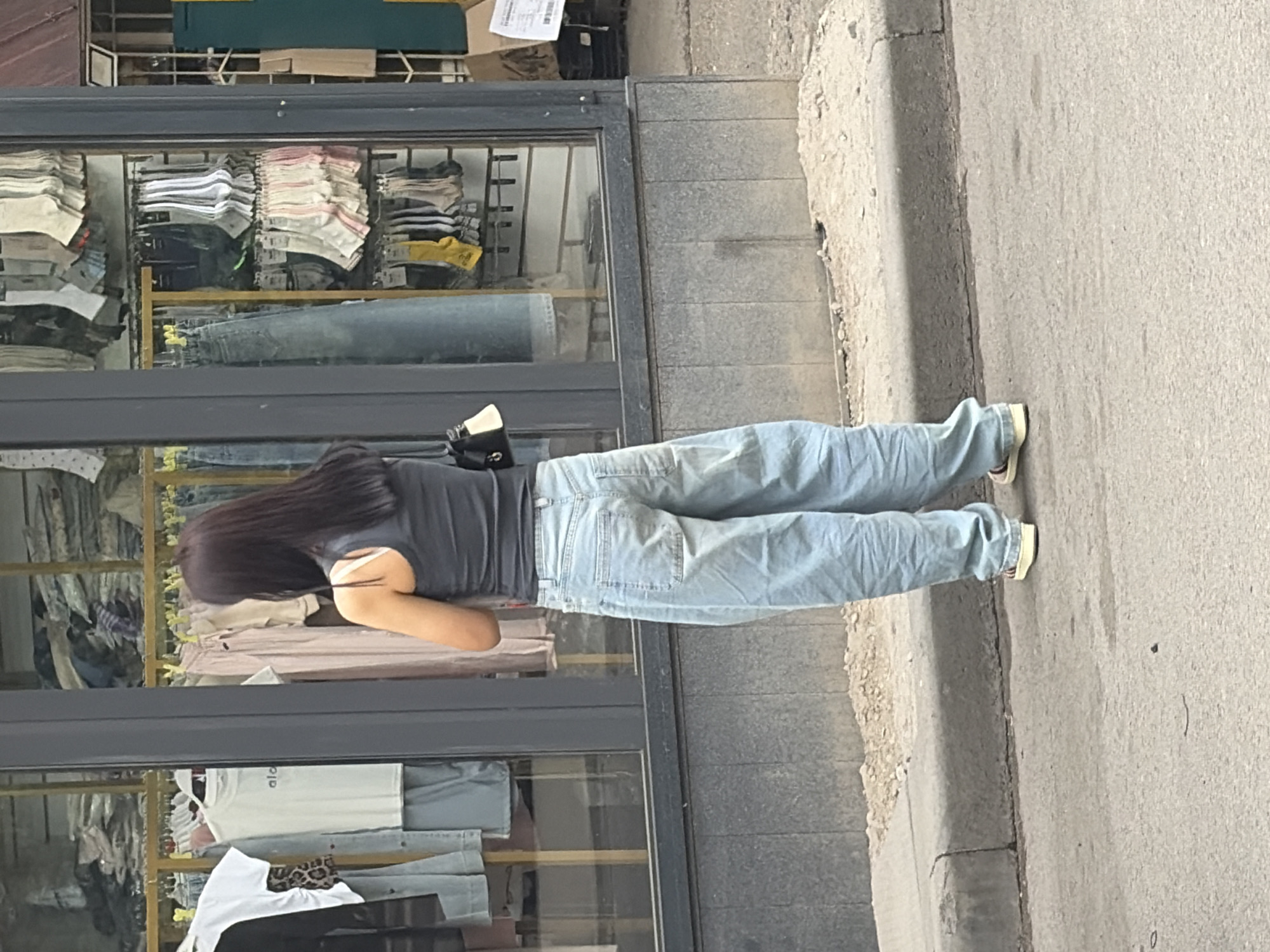 A person stands on a paved road in front of a store window displaying clothing. The person, viewed from the back, wears a dark sleeveless top and light blue, baggy jeans. They have long, dark hair. The store window shows racks of clothing, including shirts and jeans. The ground in front of the store is a mix of pavement and gravel. The scene appears to be outdoors in an urban setting.