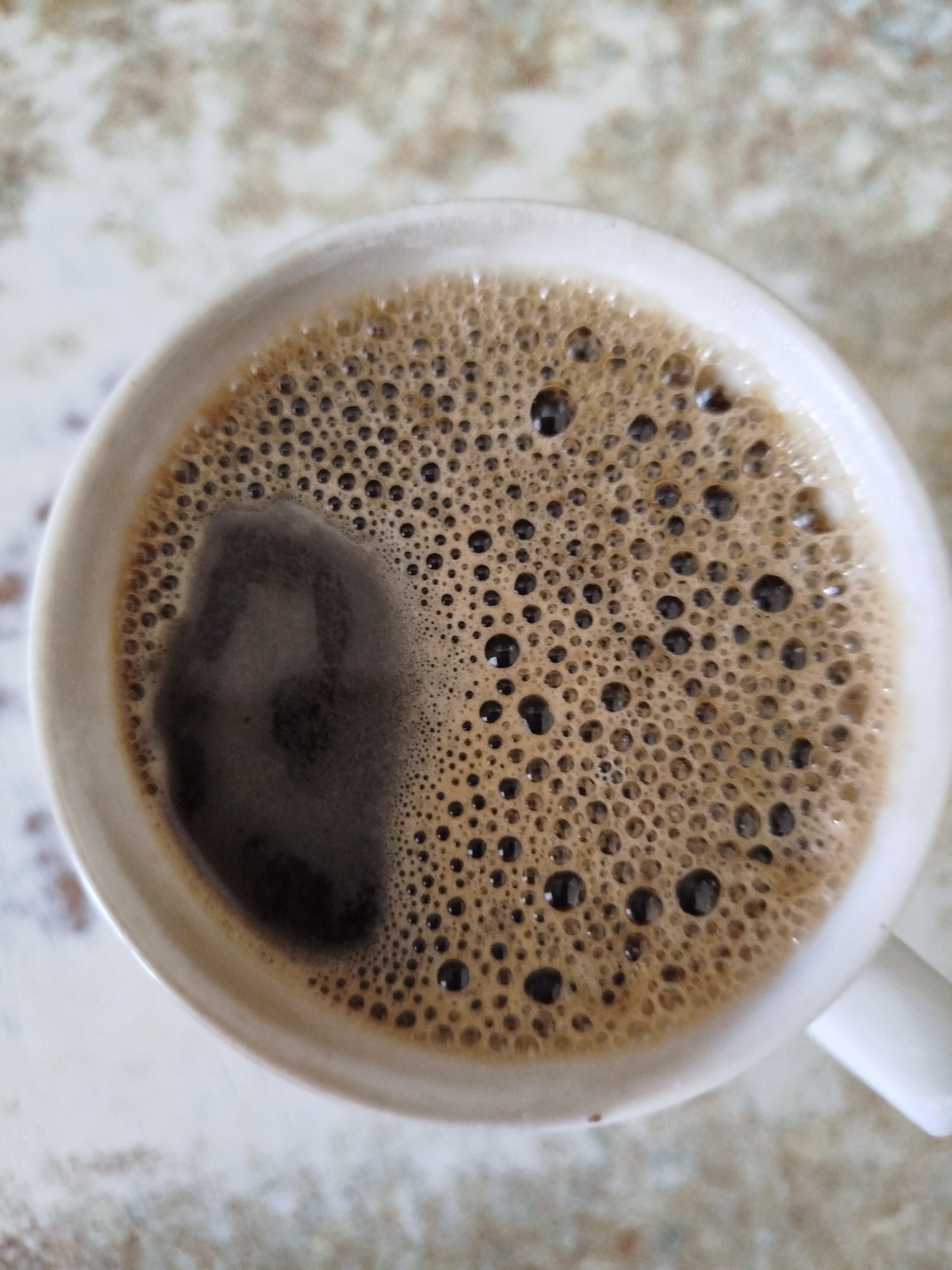 The image shows a close-up overhead view of a white mug filled with dark coffee, topped with a layer of foam. The foam consists of numerous small, uniformly sized bubbles, with a slightly darker, larger patch of foam on the left side of the mug. The coffee mug is placed on a light-colored surface with a speckled or marbled pattern. The background is out of focus. There are no people visible in the image. The location in Крывасельскі сельскі Савет, Belarus, is not depicted or suggested by any visual elements in the image.