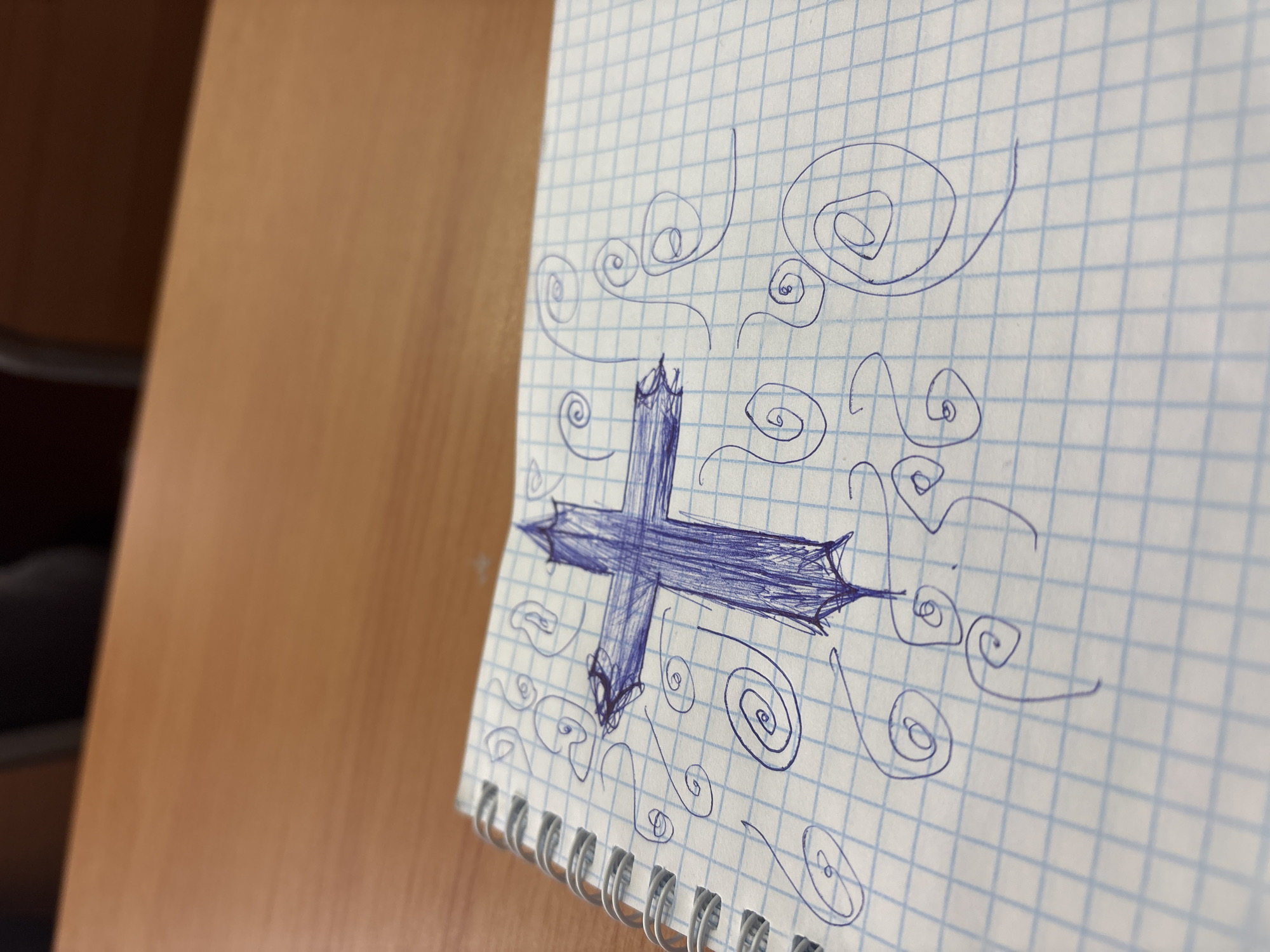The image displays a close-up, angled view of an open, spiral-bound notebook resting on a wooden surface. The notebook paper features a light blue grid pattern. The central focus of the image is a drawing rendered in dark blue ink on the notebook page.
The drawing depicts a prominent cross-like shape, thick and filled in with numerous pen strokes, creating a sense of shading. The ends of the cross's arms are stylized with sharp, pointed, almost jagged edges. Surrounding this central cross are various freehand spirals and wavy lines, also drawn in blue ink, filling the rest of the visible paper surface as a decorative or abstract pattern.
The notebook's white spiral binding is visible along the left edge of the page. To the left and bottom of the notebook, a warm, light brown wooden surface, likely a desk or table, is visible, with its grain texture apparent. The perspective of the photograph creates a shallow depth of field, with the background of the wooden surface becoming increasingly blurred towards the left. The lighting is bright and even, suggesting an indoor setting during the daytime. No discernible text is visible in the image.