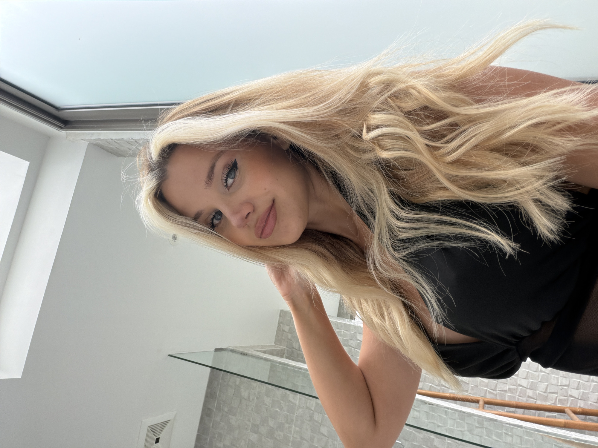 A young Caucasian female with blonde hair and blue eyes is taking a selfie in a bathroom. She is wearing a black dress with a plunging neckline. Her left arm is raised and her hand is resting on the back of her head. The background shows a tiled wall and a frosted glass partition, typical of a modern bathroom. The lighting appears to be natural, coming from a window or skylight. The city of Thep Krasatti, Thailand is not visible or implied in this image.