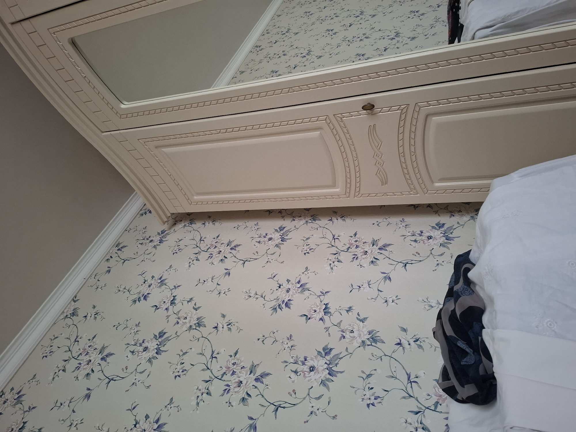 A light-colored wardrobe with mirrored doors stands in a room with floral wallpaper. A portion of a bed with white bedding is visible in the foreground, with a folded dark-colored garment placed on it. The scene appears to be an indoor domestic setting.