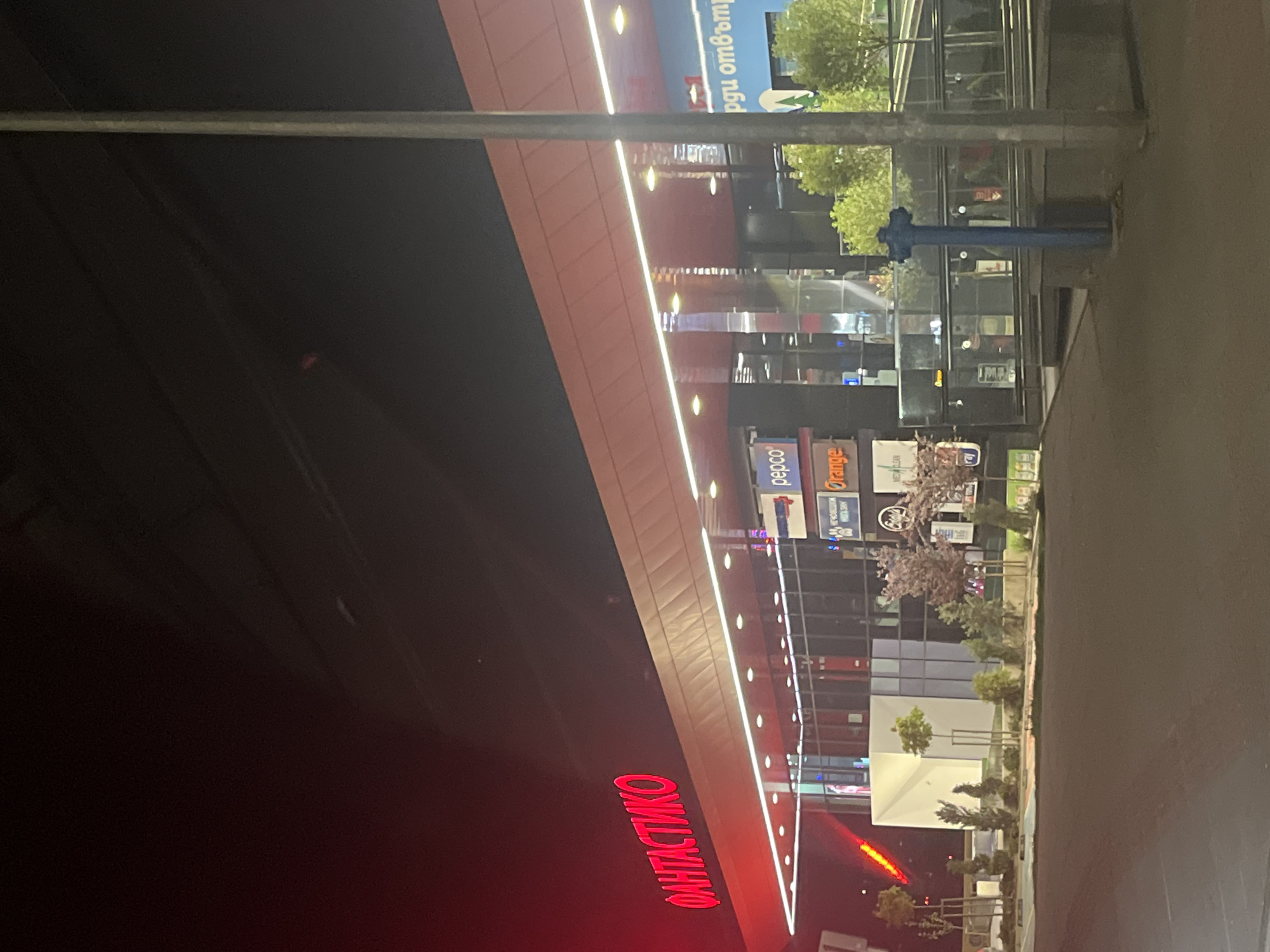 The image depicts the exterior of a commercial building at night in Sofia, Bulgaria. The word "ФАНТАСТИКО" ("FANTASTICO") in red neon letters is prominently displayed on the building's facade. Storefront signs include "pepco" and "Orange". The building is illuminated with linear white lights. Trees and shrubs are along the sidewalk, which is visible. A utility pole is in the center of the frame. The scene is likely urban.