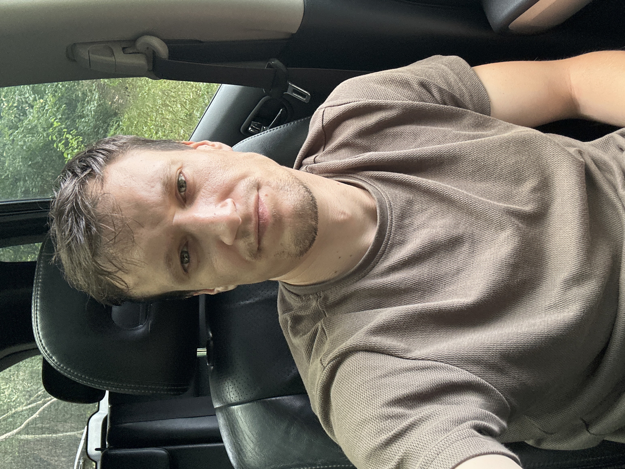 A middle-aged man with light skin, brown hair, and light-colored eyes is taking a selfie inside a vehicle. He is wearing a taupe-colored t-shirt. The man has stubble on his chin. The interior of the vehicle is visible behind him, including black leather seats and a seatbelt. Outside the car window, there is greenery, suggesting a natural or rural setting. The image appears to be taken during the daytime. The location in Makiv, Ukraine is not discernible from the image itself.