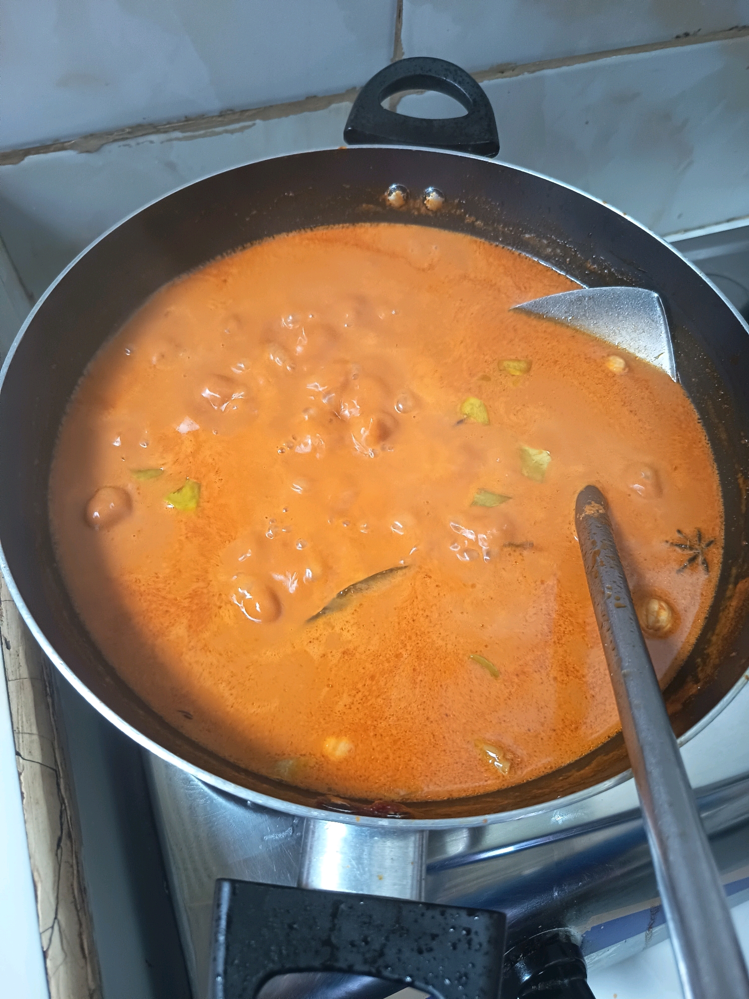 In the city of Aluekumba, Indonesia, a close-up, overhead shot captures a pan of simmering curry on a stovetop. The curry, a vibrant orange hue, is smooth with a few visible ingredients, including pieces of protein and perhaps some aromatics like ginger or garlic. A metal spatula rests in the pan, its handle extending out of frame. The pan itself is dark-colored with a silver rim, and its handle is black. The background reveals a tiled kitchen wall, suggesting a domestic setting. The stovetop is visible at the bottom, with a portion of the burners and control knobs. The lighting is ambient, highlighting the rich texture and color of the curry.