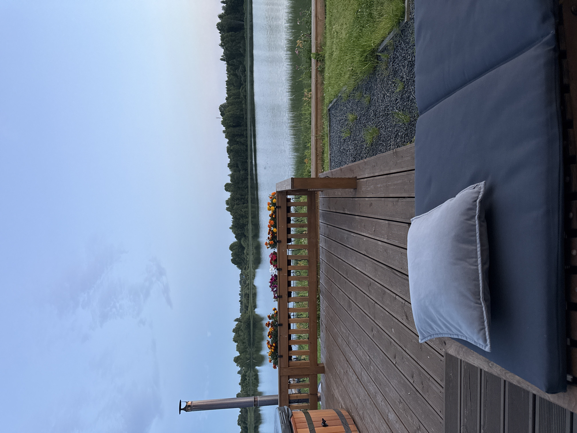 The image depicts an outdoor wooden deck overlooking a tranquil body of water, likely a lake, under a pale blue twilight sky. The deck features a wooden railing adorned with flower boxes filled with vibrant orange and pink flowers. To the left, a portion of a wooden hot tub is visible, along with a tall metal chimney. In the foreground, a dark grey chaise lounge is set with a light grey cushion. The water is calm, reflecting the soft hues of the sky and the distant tree line that borders the far shore. Tall grasses and some smaller flowering plants grow along the water's edge. The overall scene conveys a sense of peaceful relaxation in a natural setting, possibly a lakeside retreat or a residential property. No individuals are present in the frame, and no specific landmarks indicating a precise location in Pyhäjärvi, Russia, are discernible.