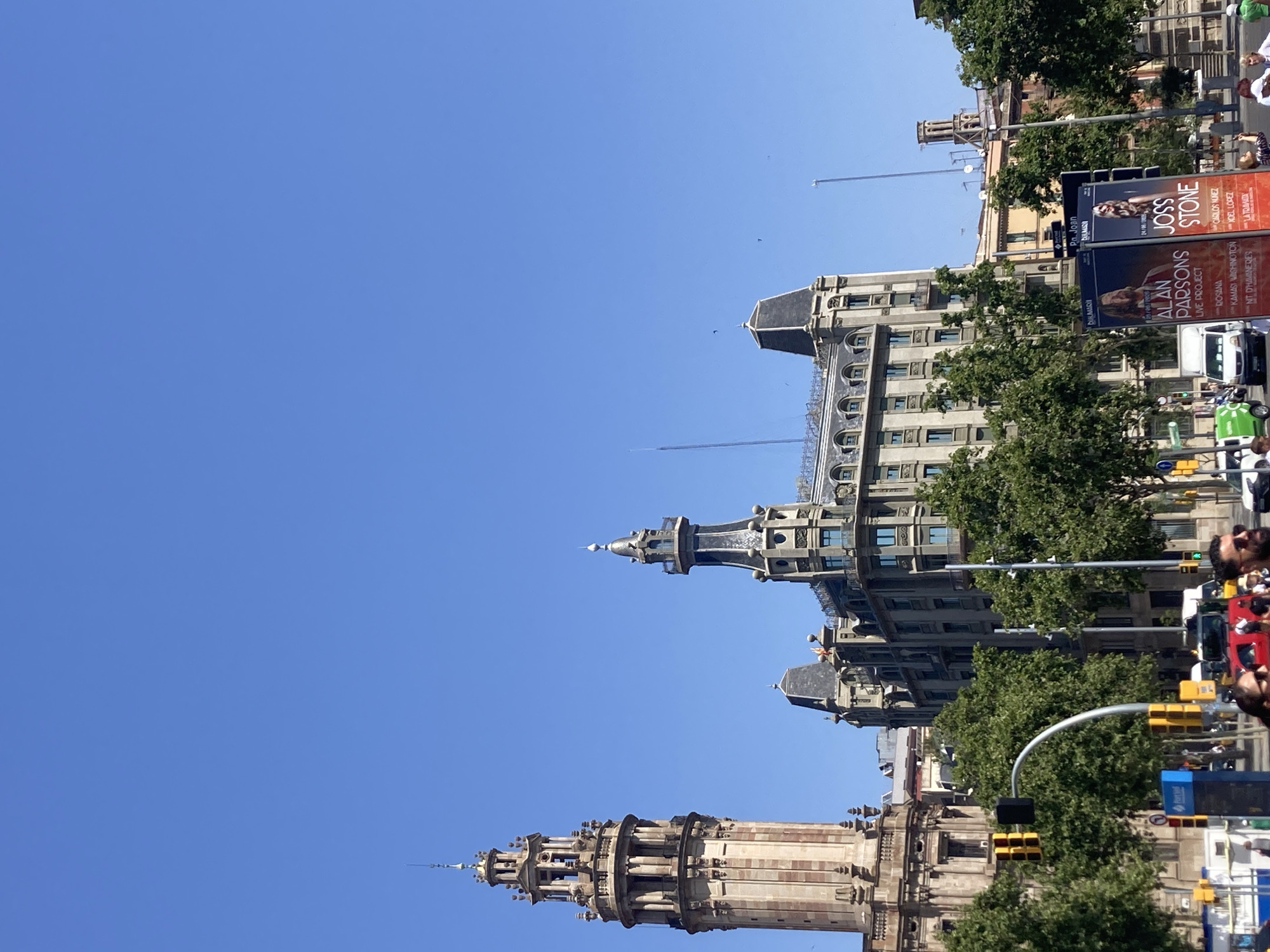 This is a vertical-format, eye-level shot of a street scene in Barcelona, Spain, under a clear, bright blue sky. The image is dominated by the architecture of several ornate buildings, with intricate stonework and tiered roofs. Two prominent towers with pointed spires are visible, one in the foreground on the lower left and another further back on the upper right, suggesting a historic or ecclesiastical area of the city. Lush green trees line the street, providing a contrast to the stone structures and the bright sky.
In the mid-ground, several billboards are displayed. One advertises "ALAN PARSONS LIVE PROJECT," and another features "JOSS STONE." Other names and phrases in Spanish and English are visible, including "ROSANA," "CARLOS NUNEZ," and "LA TRAMATA," indicating upcoming performances or events. A white van and parts of a blue bus are visible, along with traffic lights and street signs, suggesting an active urban environment.
Several people are present, though their faces are not clearly discernible due to the distance and vertical orientation. They appear to be pedestrians and possibly event attendees. The overall impression is a sunny day in a vibrant European city, likely during daytime given the bright light and clear sky. The composition, with the buildings and towers reaching upwards towards the sky, gives a sense of grandeur and scale.