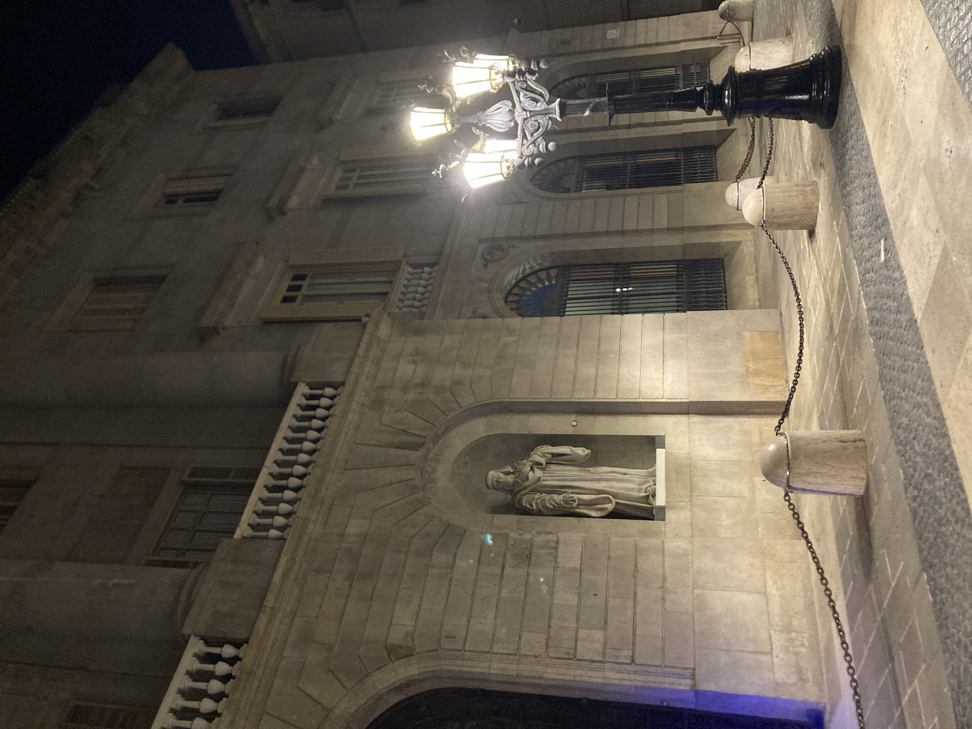 A statue of a robed figure stands in an alcove on the facade of a stone building at night. The building has multiple windows with shutters and a balustrade across the second story. A multi-headed streetlamp illuminates the foreground. The street is paved with stone and bordered by a chain fence supported by stone bollards. The scene is located on Las Ramblas in Barcelona, Spain.