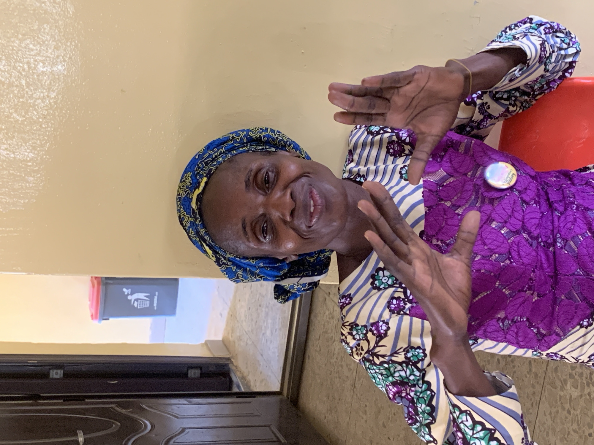 A medium shot depicts an older Black woman indoors, smiling and gesturing with her hands. She is wearing a colorful patterned headscarf and a purple lace-over-striped-fabric dress with a button on the chest. She sits in a room with a light yellow wall. Behind her and to the left, a glimpse through a doorway reveals a trash bin and tiled flooring. The location is likely in Apata Yakuba, Nigeria, given the prompt.