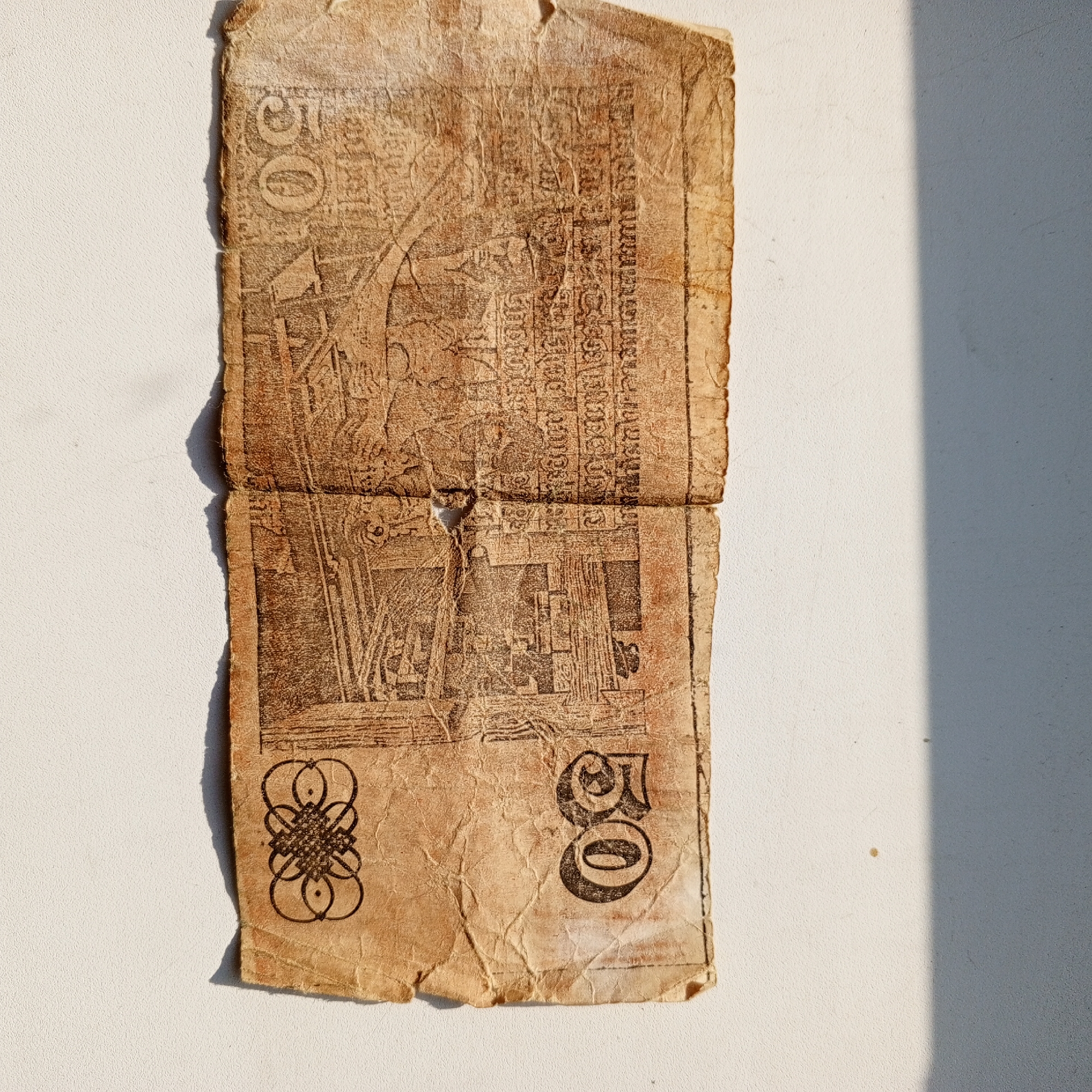 The image shows a crumpled, aged banknote with a numerical denomination of "50" visible in two locations. The banknote features intricate illustrations and text that appear to be in a script other than Latin. One side of the banknote displays a design with what looks like a stylized knot or emblem. The banknote is placed on a white, textured surface, with a shadow falling across the right side of the image, suggesting outdoor lighting. There is no visible context or information suggesting this image is related to Sayanogorsk, Russia.