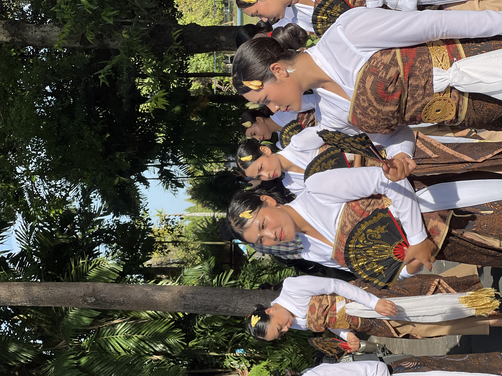 A group of women dressed in traditional Balinese attire are standing outdoors, possibly in Denpasar, Indonesia. They are wearing white long-sleeved blouses and patterned wrap skirts with ornate gold belts. Many of the women are holding open black and gold fans. Their hair is styled in buns, adorned with yellow flowers. The background consists of trees with lush green foliage and a glimpse of a body of water. The lighting suggests it is daytime, with sunlight filtering through the leaves.