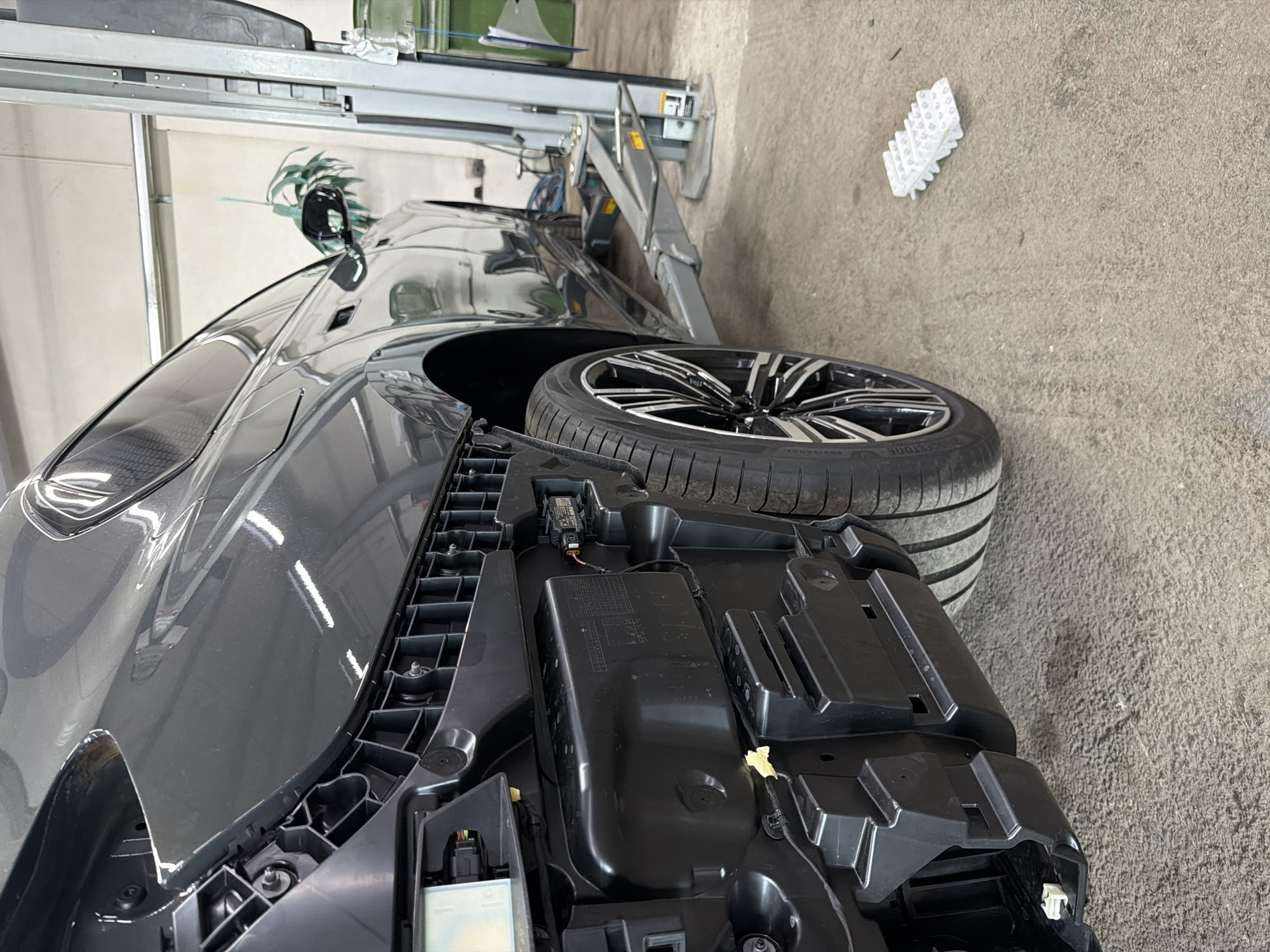 An indoor scene in an automotive workshop in Burgas, Bulgaria, features a dark grey car undergoing maintenance or repair. The car, possibly a coupe or sedan, is positioned next to a car lift, with its right rear wheel and tire prominent in the foreground. The tire clearly displays the brand name "BRIDGESTONE". A large, black plastic component, likely a part of the car's bumper or fascia, is detached and lying on the concrete floor next to the vehicle, revealing internal structural elements and some wiring.
The workshop environment is utilitarian, with light-colored walls and a textured concrete floor. In the background, the metallic structure of a car lift is visible, suggesting a professional repair setting. A small, white corrugated plastic item rests on the floor further in the background. A leafy green plant or decorative element can be seen behind the car's side mirror, adding a touch of greenery to the industrial setting. The lighting suggests daytime, likely supplemented by artificial garage lights. No people are visible in the image.
