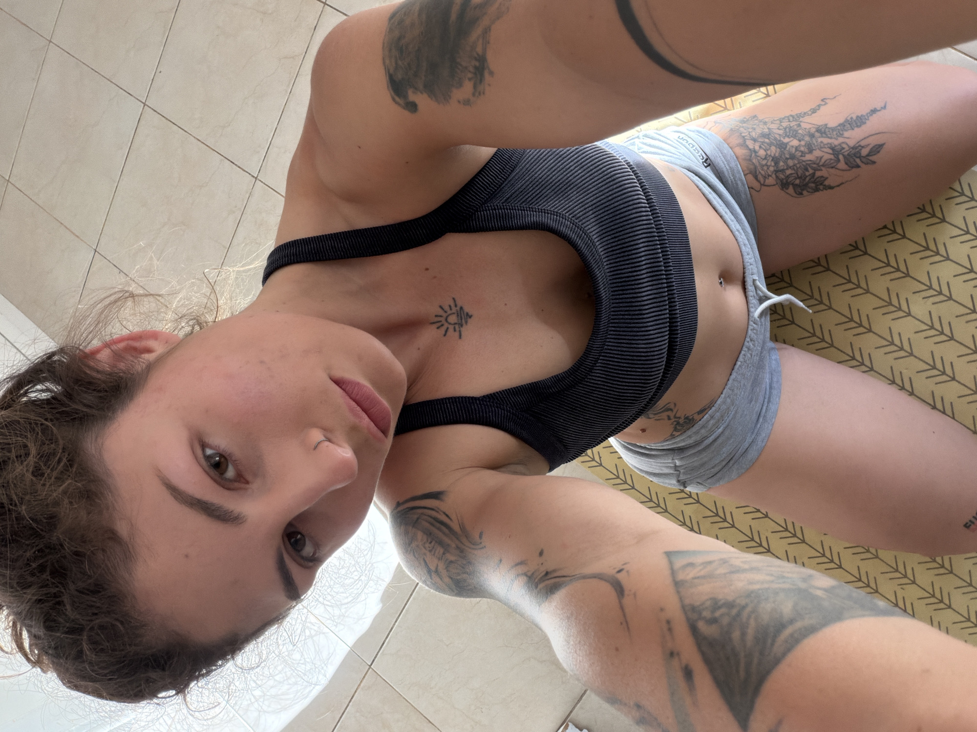 A woman lies on a patterned yoga mat in what appears to be a room with tiled flooring. She is wearing a striped black sports bra and grey shorts. She has multiple tattoos visible on her arms, chest, and thighs. Her body is oriented with her head towards the left and her legs towards the right of the frame. Her face is slightly tilted. The location in Kiryat Bialik, Israel, cannot be determined based on the provided image.