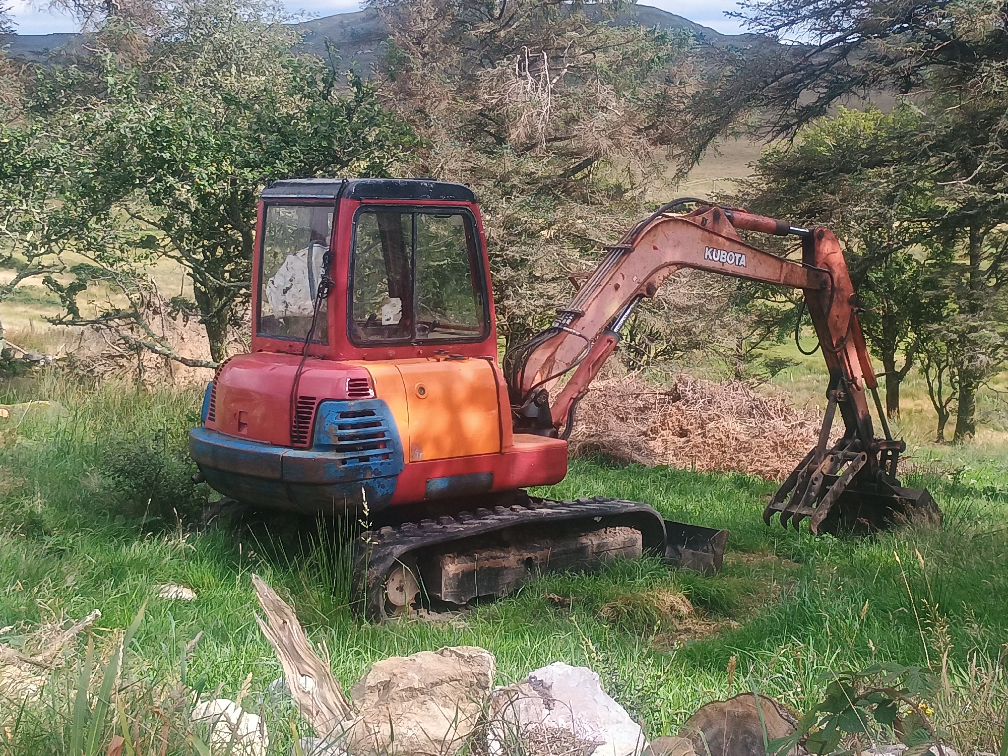 A red and orange Kubota excavator sits in a grassy field, with trees and hills in the background. The excavator is parked on a grassy slope, with its tracks partially sunk into the ground. The cab of the excavator is red, with a black roof and a large glass window. The boom and arm of the excavator are orange, and the bucket is black. The excavator appears to be old and weathered, with rust and paint chips visible on its surface. The background of the image consists of a dense forest of trees, with some areas of bare ground and rock visible. In the distance, a range of hills can be seen, with a cloudy sky overhead. The overall scene is one of rural isolation, with the excavator appearing to be abandoned or unused.