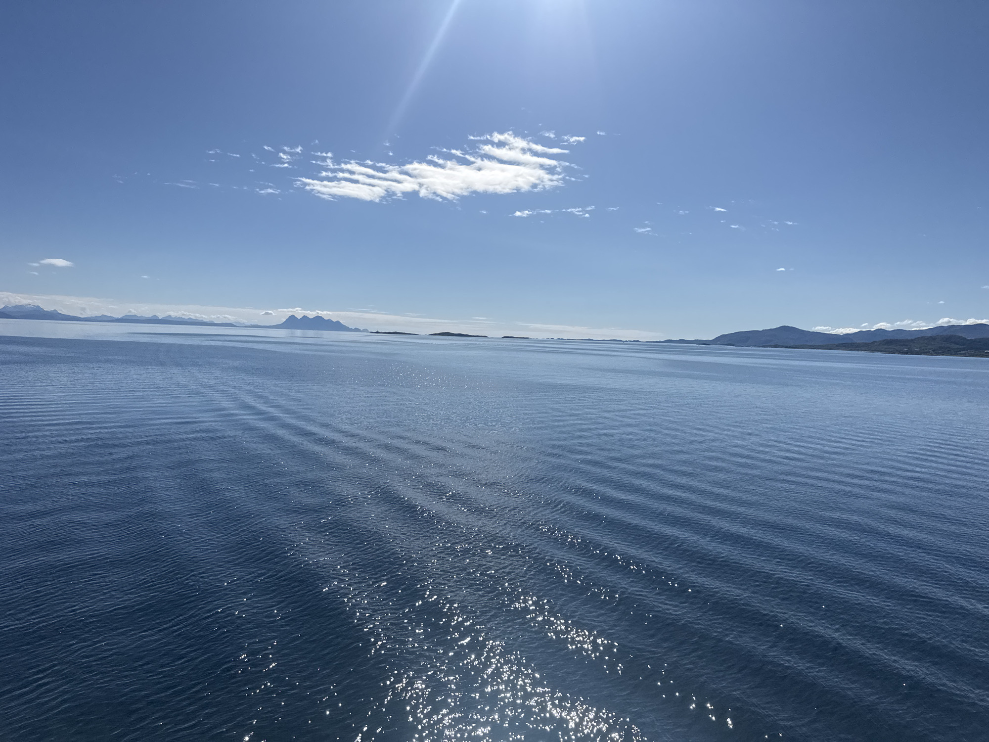 This is a photograph of a body of water, likely a fjord, with mountains in the distance. The sky is blue with a few scattered clouds. The sun is shining brightly, creating reflections on the water. The image appears to have been taken from a boat or other vessel, as there are visible ripples on the water's surface, suggesting movement. The distant landmasses are hazy and appear to be mountainous. The scene suggests a clear, sunny day in a coastal or maritime environment. The location is specified as Lødingen, Norway.